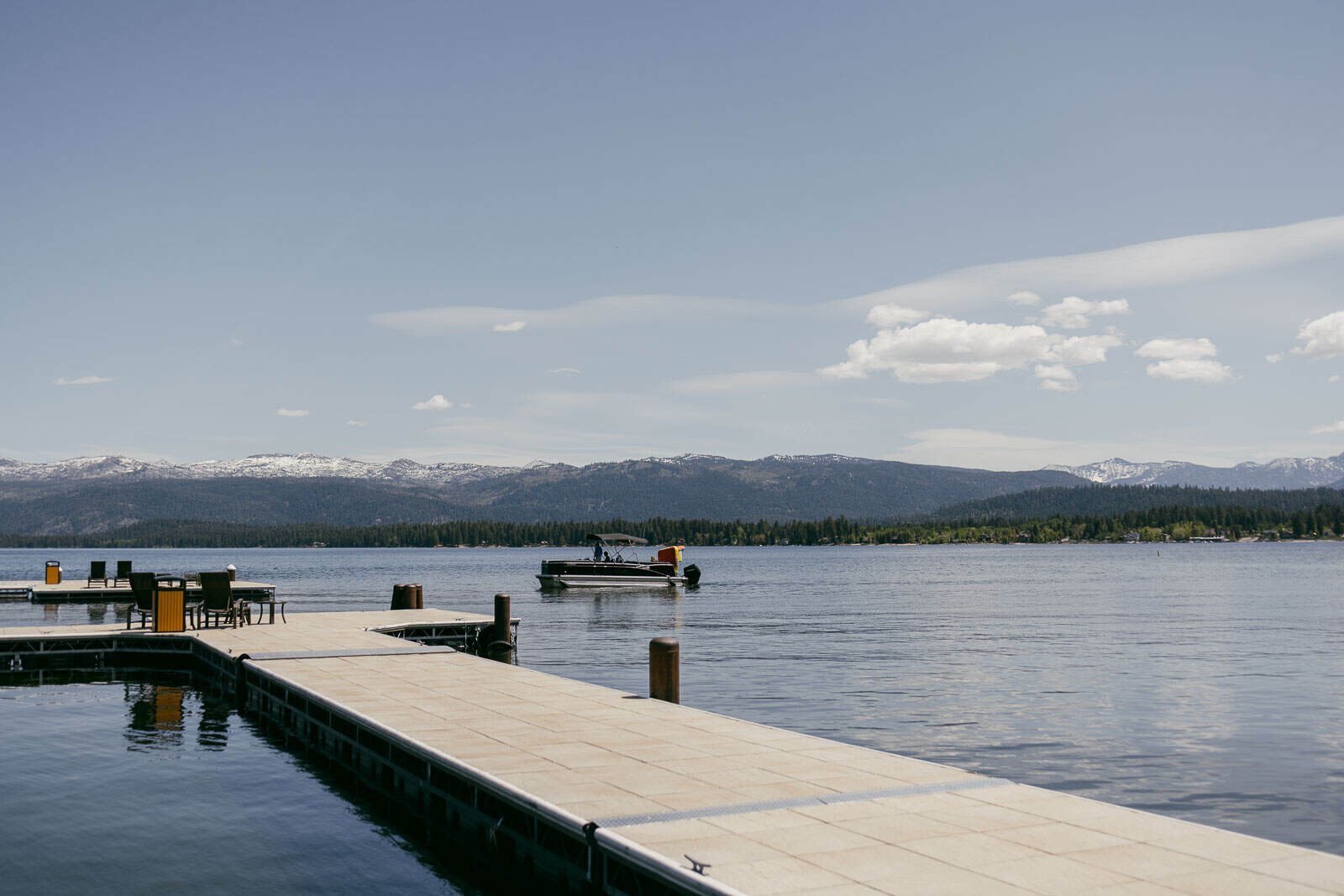  A calm Payette Lake with a dock in the foreground, a pontoon boat on the water, and distant snow-capped mountains under a partly cloudy sky. Trees line the shore across the lake. 