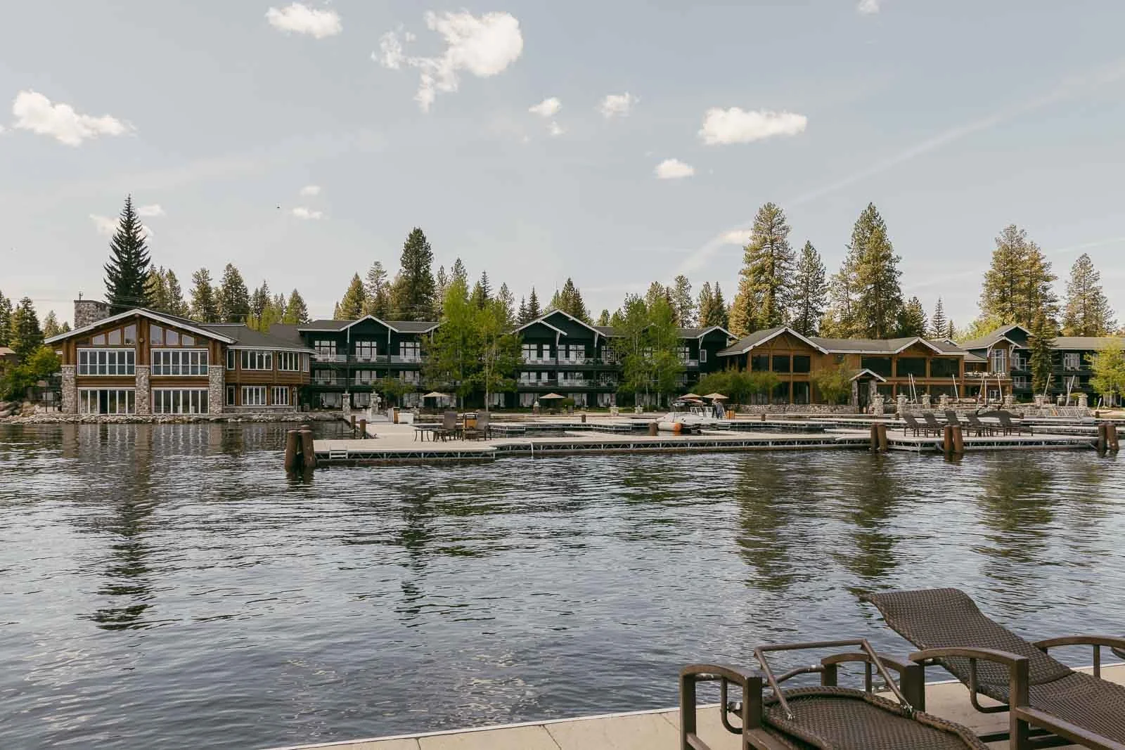 Lakeside view of Shore Lodge's wooden lodges and cabins surrounded by trees, with empty docks extending into the calm water in the foreground. Lounge chairs sit on the dock under a partly cloudy sky.