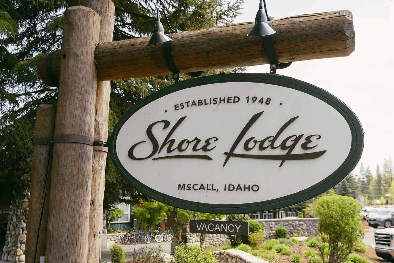  A wooden sign reads "Shore Lodge, Established 1948, McCall, Idaho" with a "Vacancy" sign below, surrounded by trees and part of a stone building in the background. 