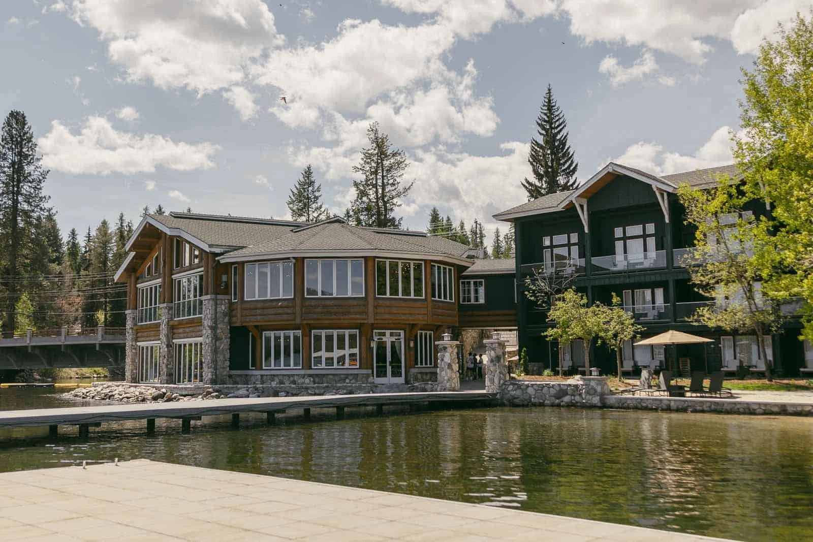  Modern lakeside buildings with large windows and wood accents stand surrounded by trees. A dock extends over calm water in the foreground under a partly cloudy sky. 