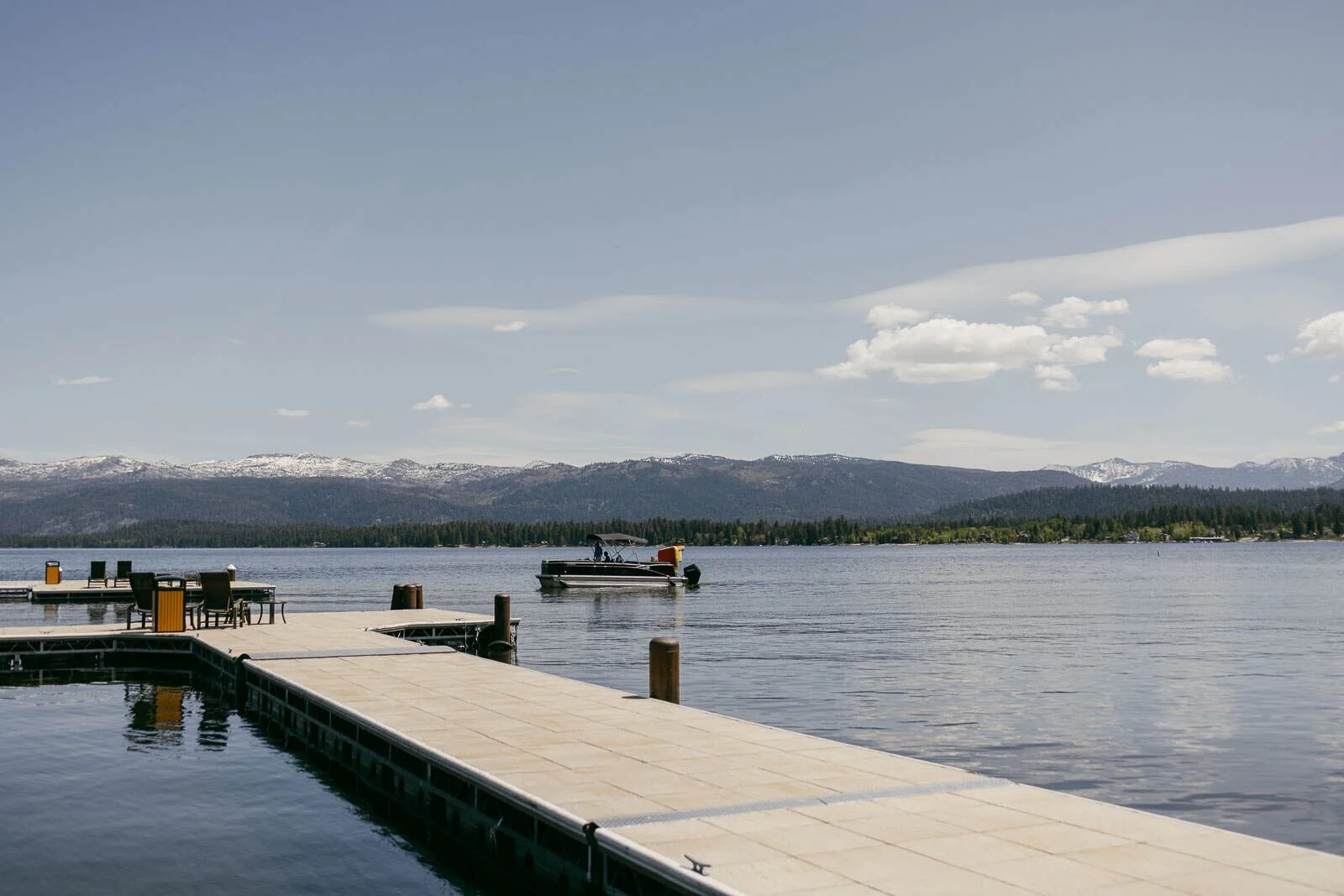  A calm lake with a dock in the foreground and a pontoon boat on the water. Trees and snow-capped mountains are visible in the distance under a clear sky. 