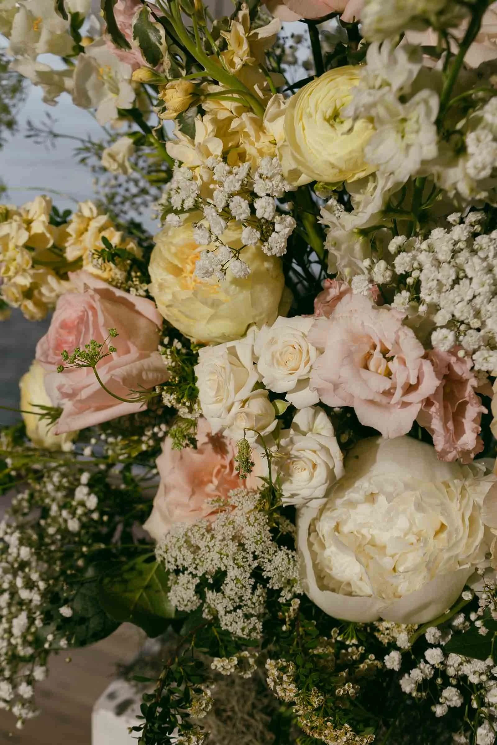  A close-up of a floral arrangement featuring pale yellow, white, and soft pink roses, peonies, and delicate white filler flowers with lush green foliage in the background. 