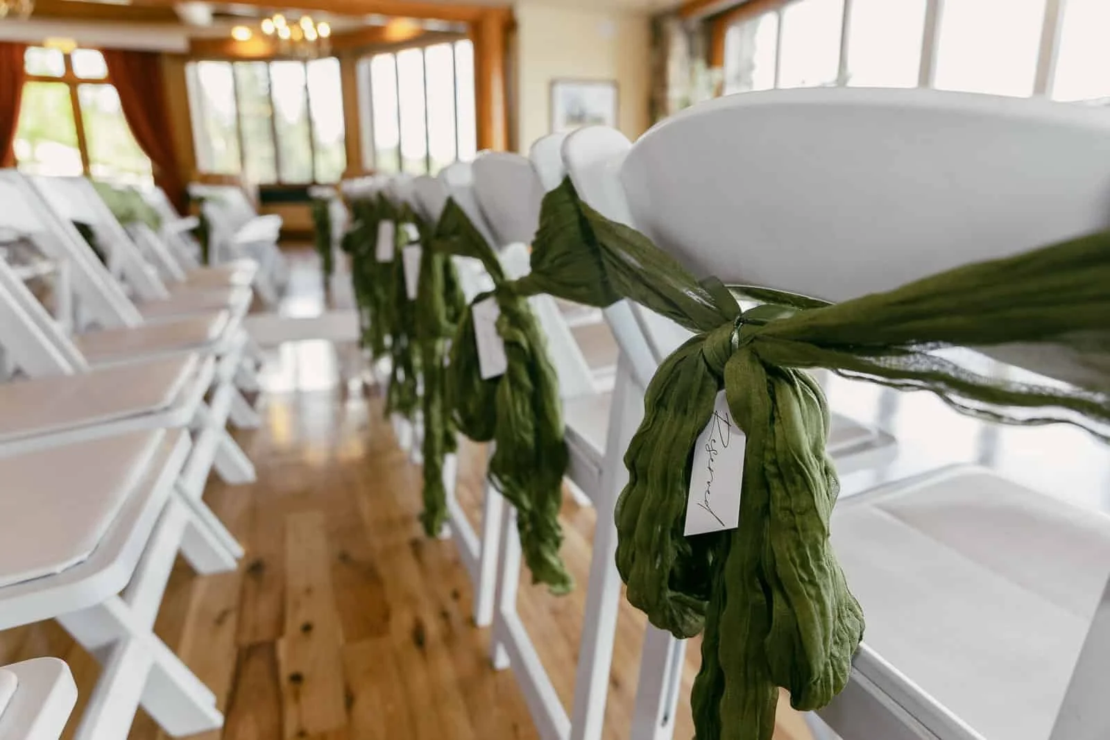  Rows of white chairs decorated with green fabric sashes and name tags, arranged indoors on a wooden floor with large windows in the background. 