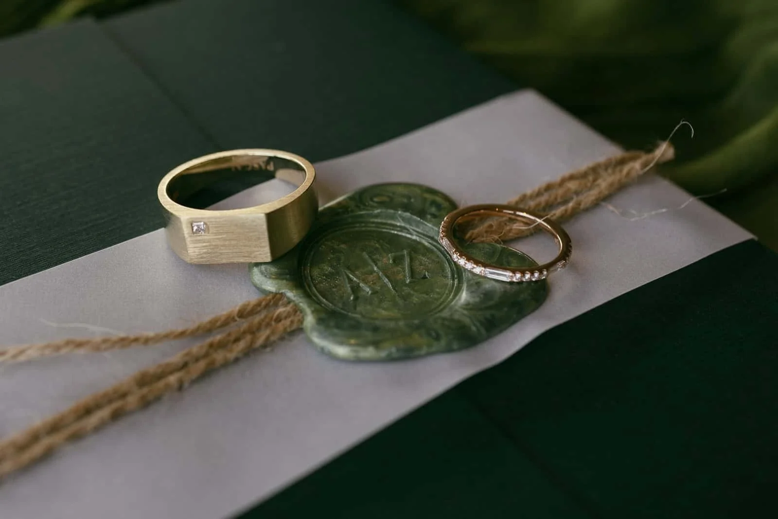  Two rings rest on a green wax seal atop a dark green envelope wrapped with twine. One ring is gold with a small diamond, and the other is a thin gold band with small stones. 