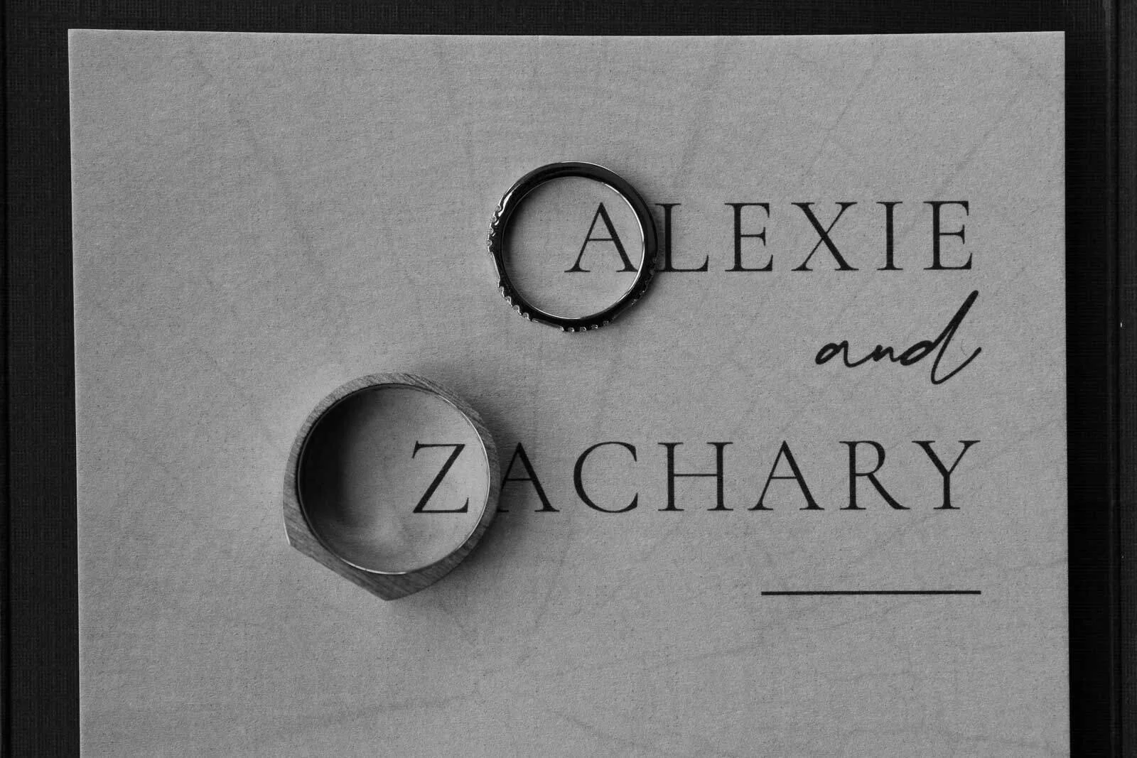  Two rings rest on a card with the names “Alexie and Zachary” printed in large letters. The card has a subtle textured background and the photo is in black and white. 