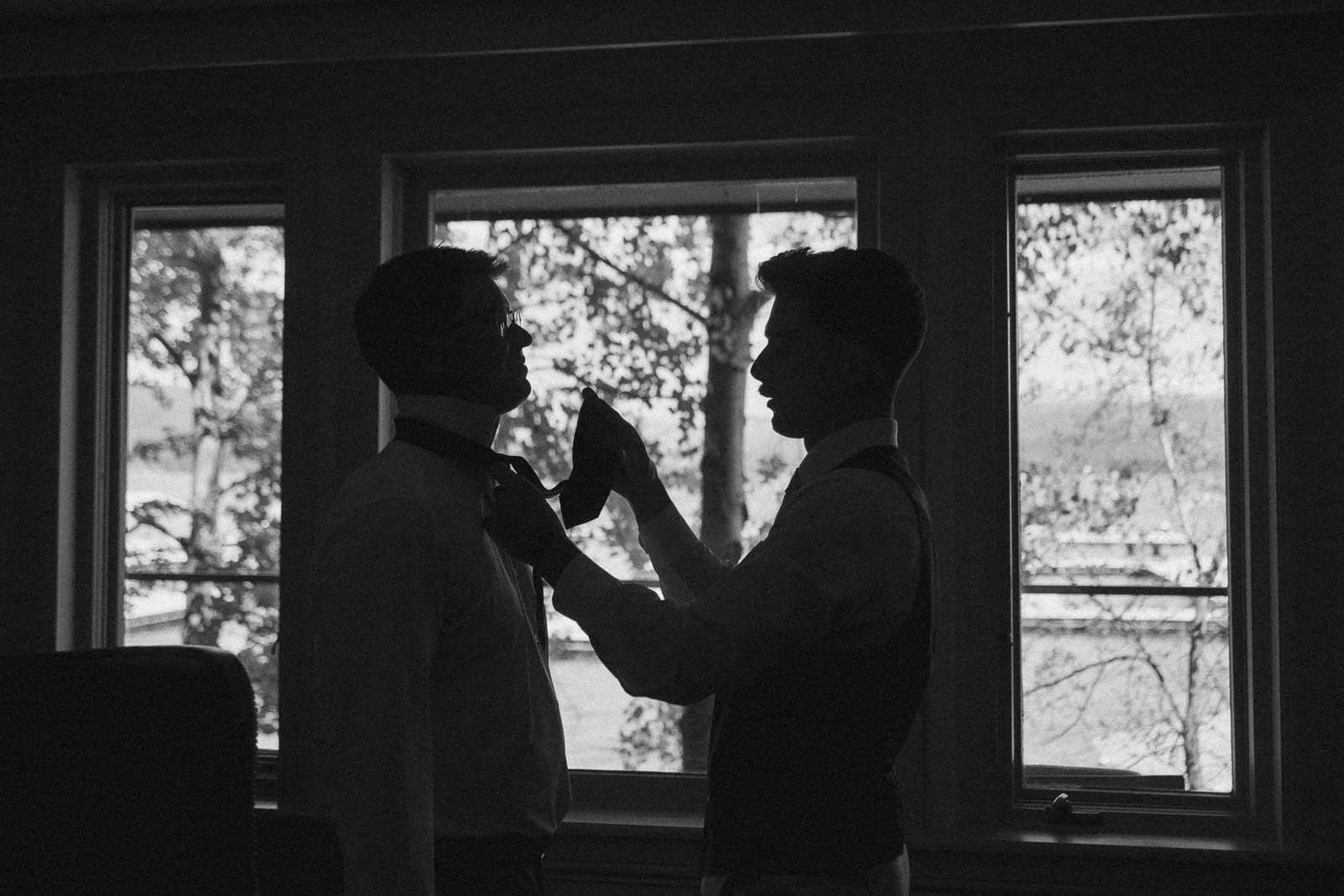  Two people in silhouette stand by large windows; one adjusts the other's tie. Trees and a body of water are visible outside, in black and white. 