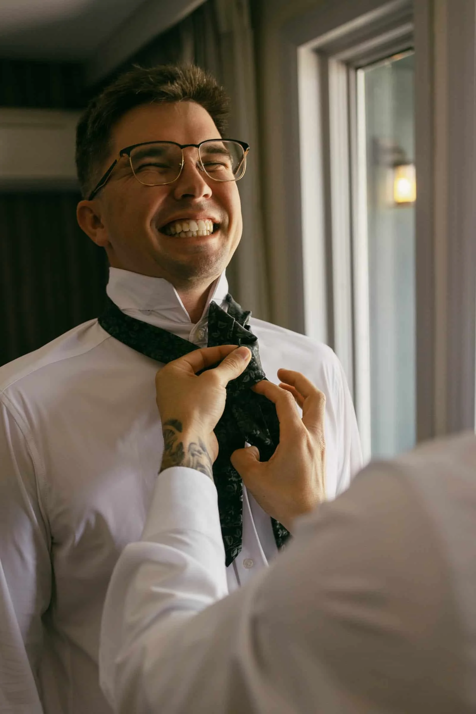  A smiling groom in glasses is getting their tie adjusted by another person, standing indoors by a window. 