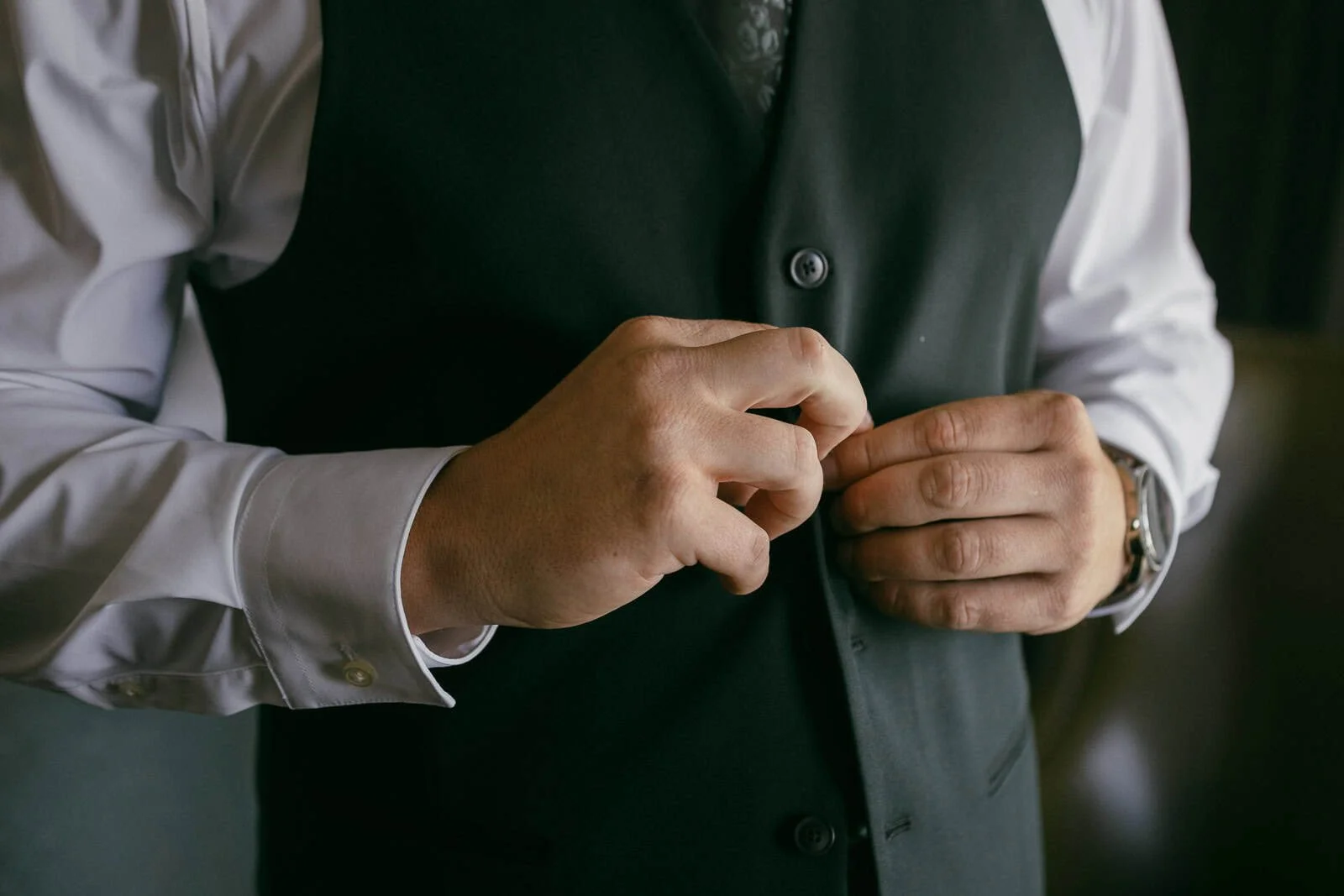  A groom wearing a white shirt and dark vest is buttoning their vest. Only their upper body and hands are visible. 