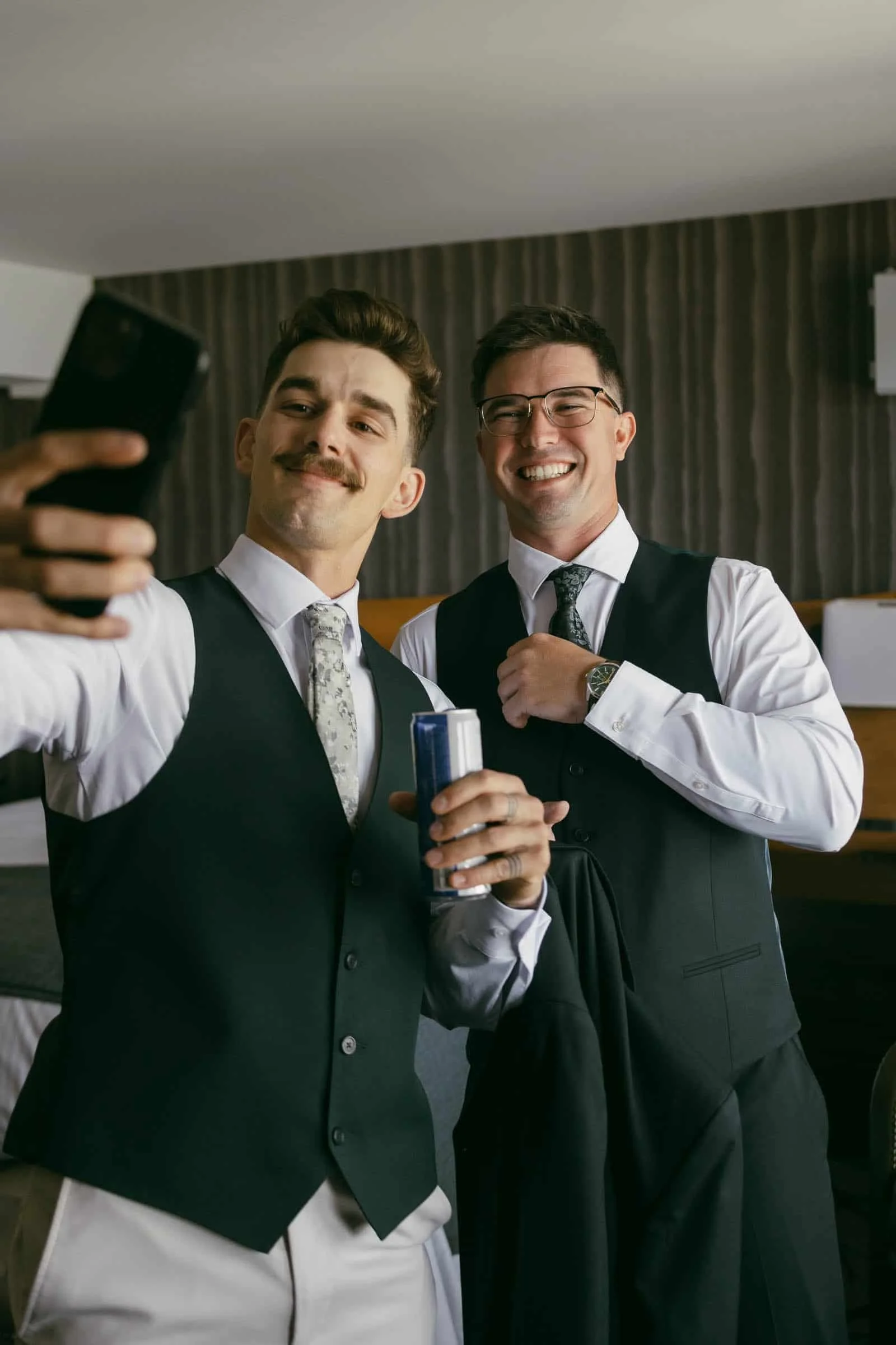  Two men in suits smile for a selfie; one holds a phone and a can, while the other adjusts his tie. They appear to be dressed for a formal event, standing indoors. 