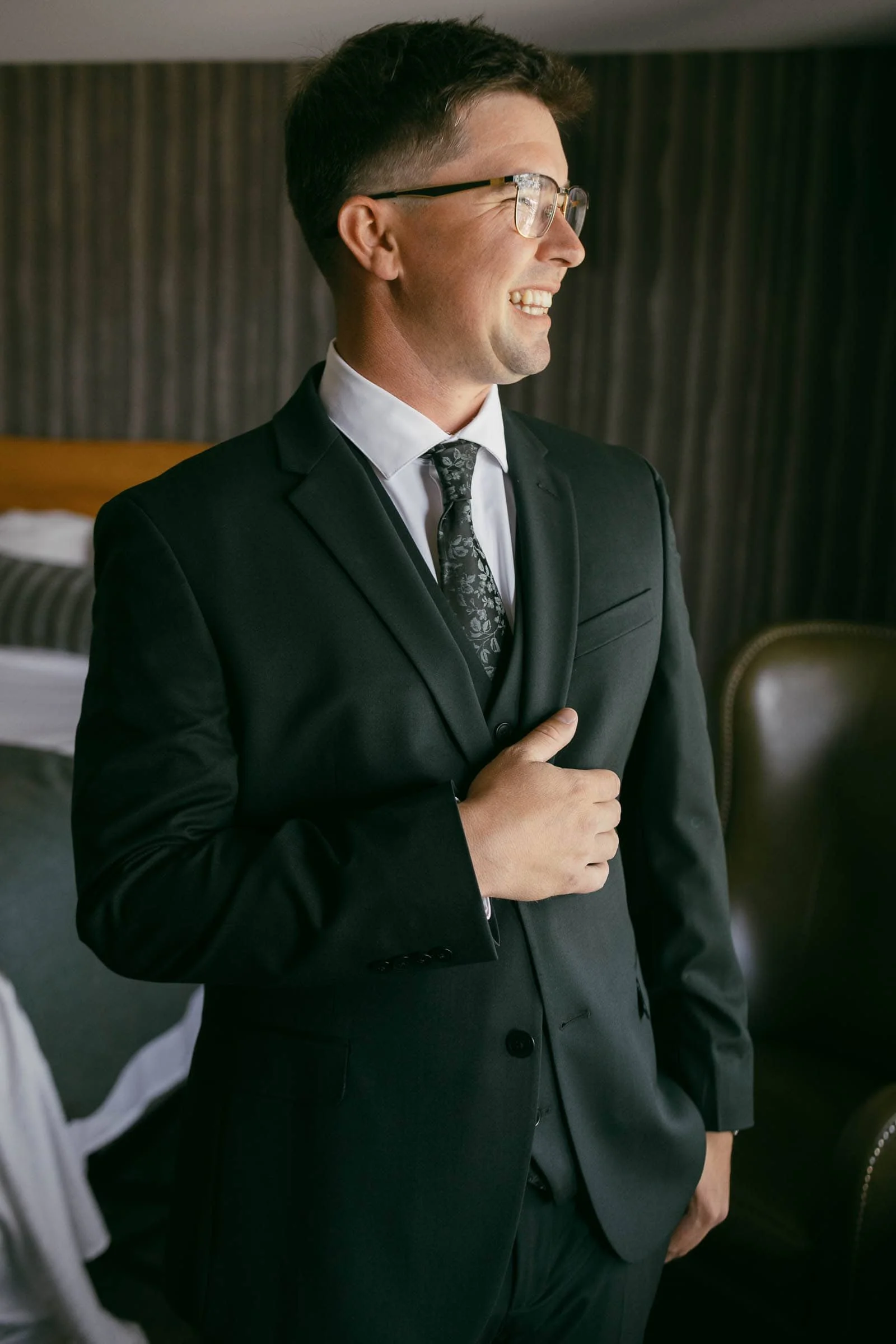  A man wearing glasses and a dark suit smiles while standing indoors near a bed and a chair, with one hand adjusting his jacket. 