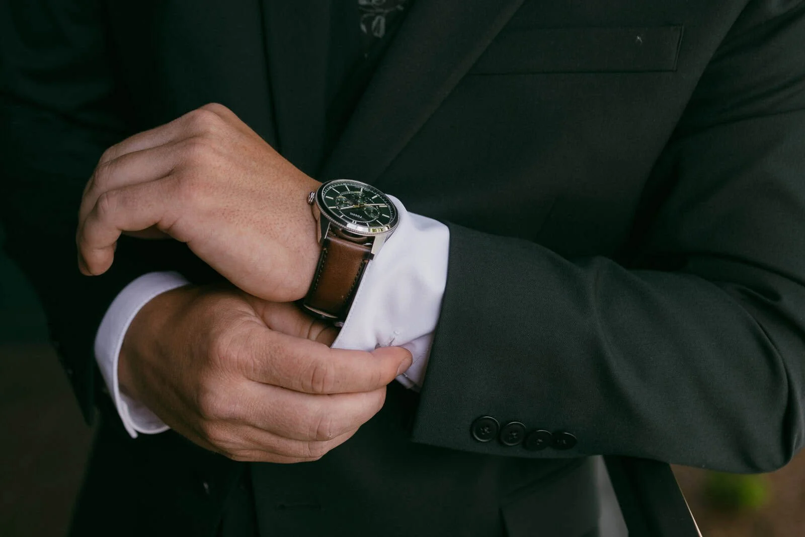  A person in a dark suit adjusts their brown leather wristwatch, with one hand holding the watch and the other pulling back a white shirt cuff. 