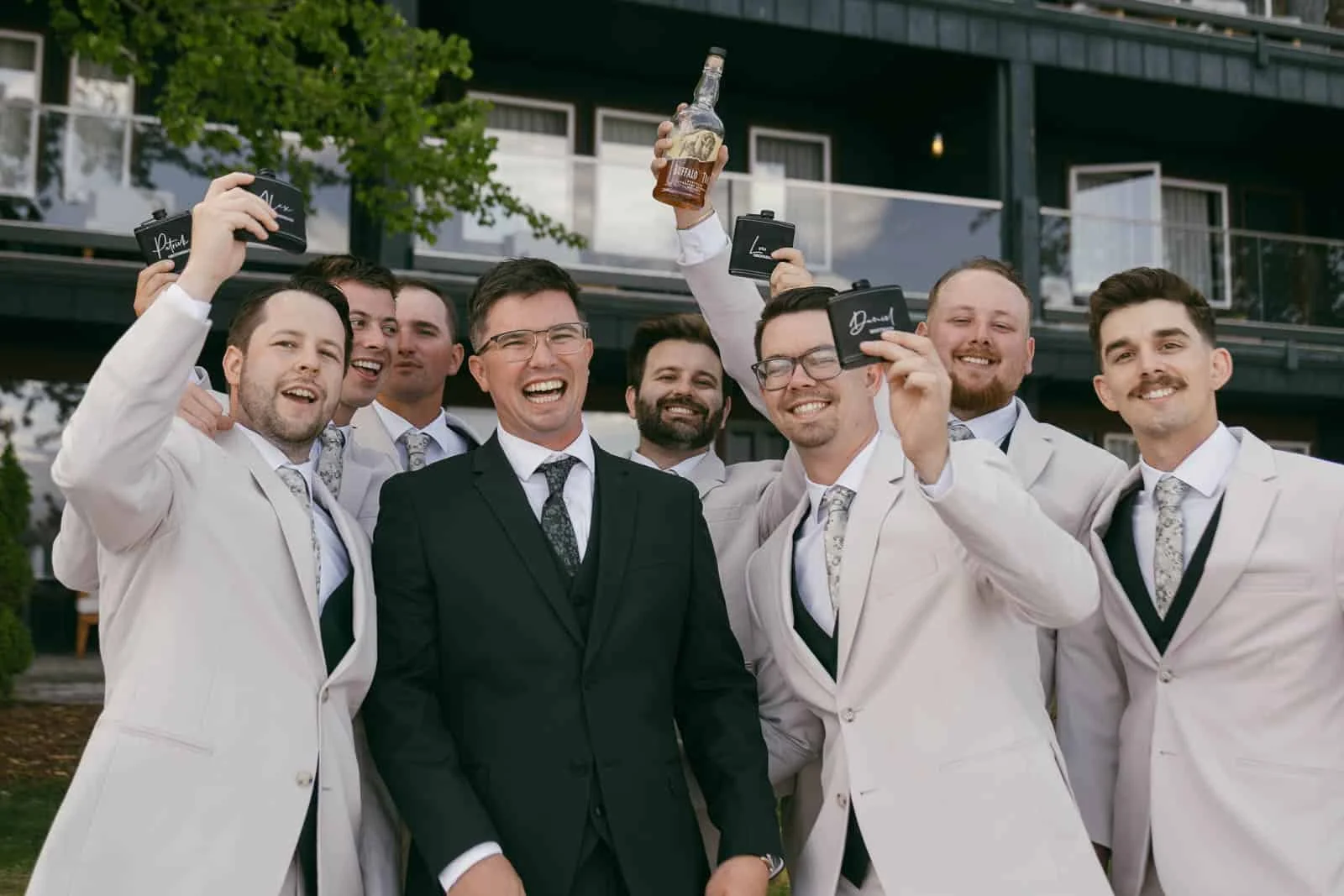  A group of men in suits, with one in a black suit and others in light suits, smile and pose outdoors. Some hold flasks; one holds up a bottle of whiskey. 