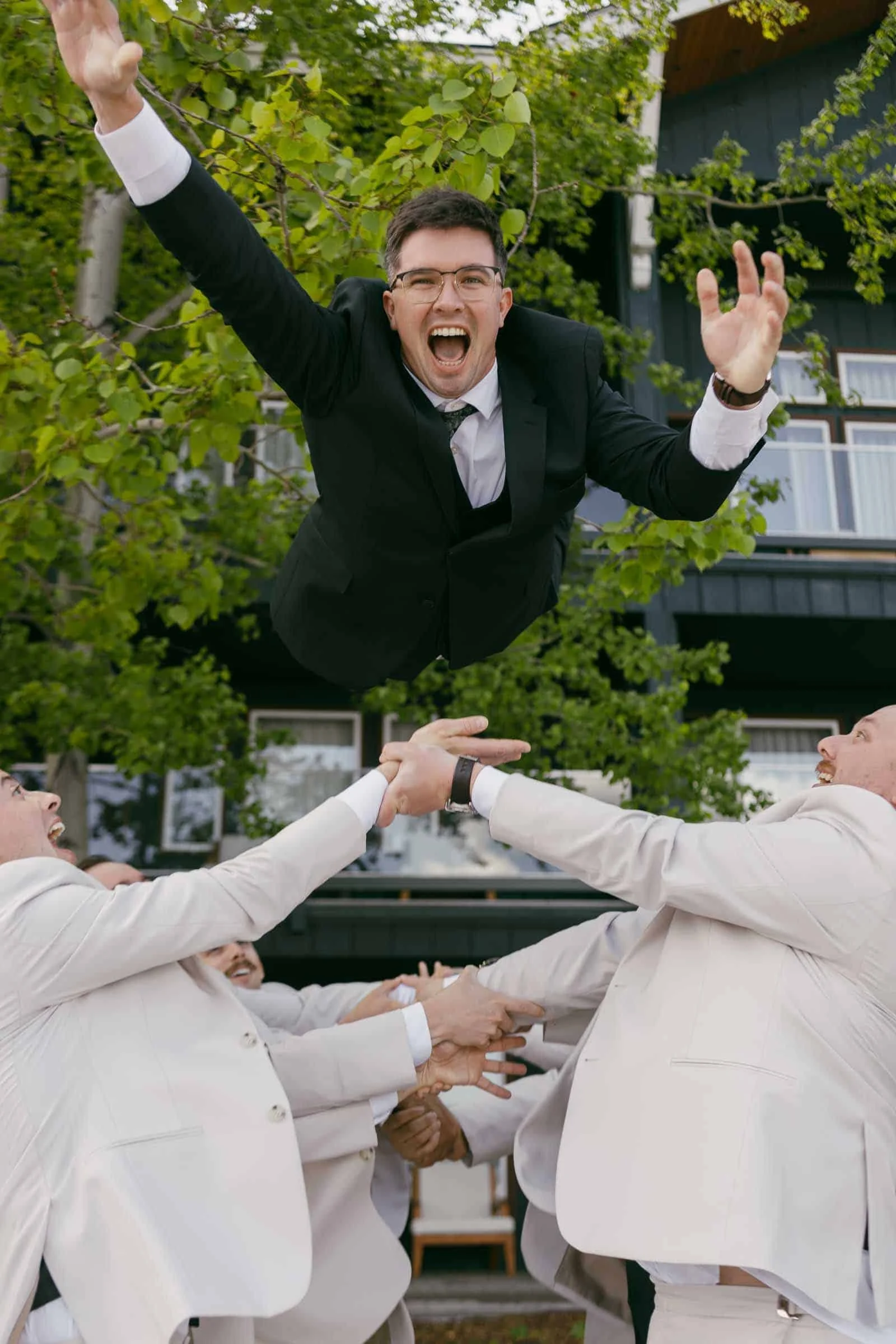  A joyful groom in a green suit is tossed into the air by a group of people in white suits, smiling and celebrating outdoors near a building with green trees in the background. 