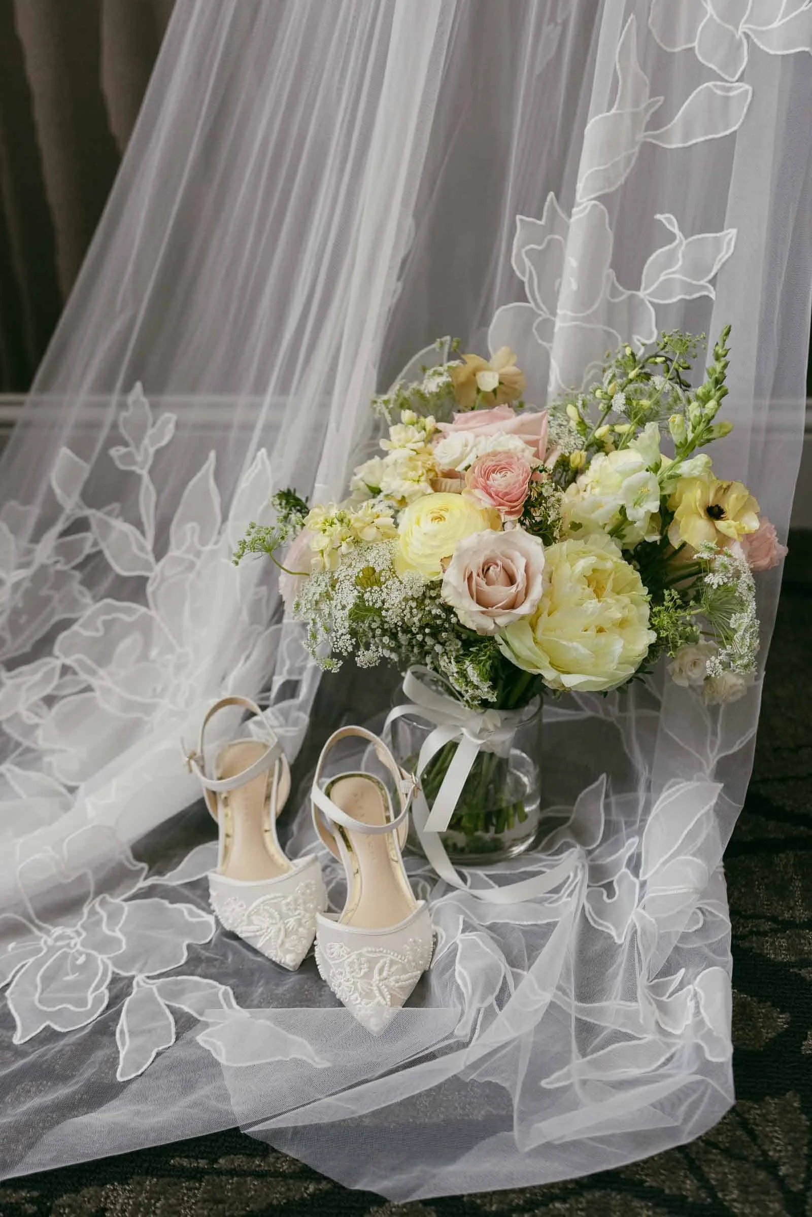  Bridal shoes and a pastel bouquet of roses and greenery rest on floral embroidered tulle, likely part of a wedding dress or veil, on a dark floor. 