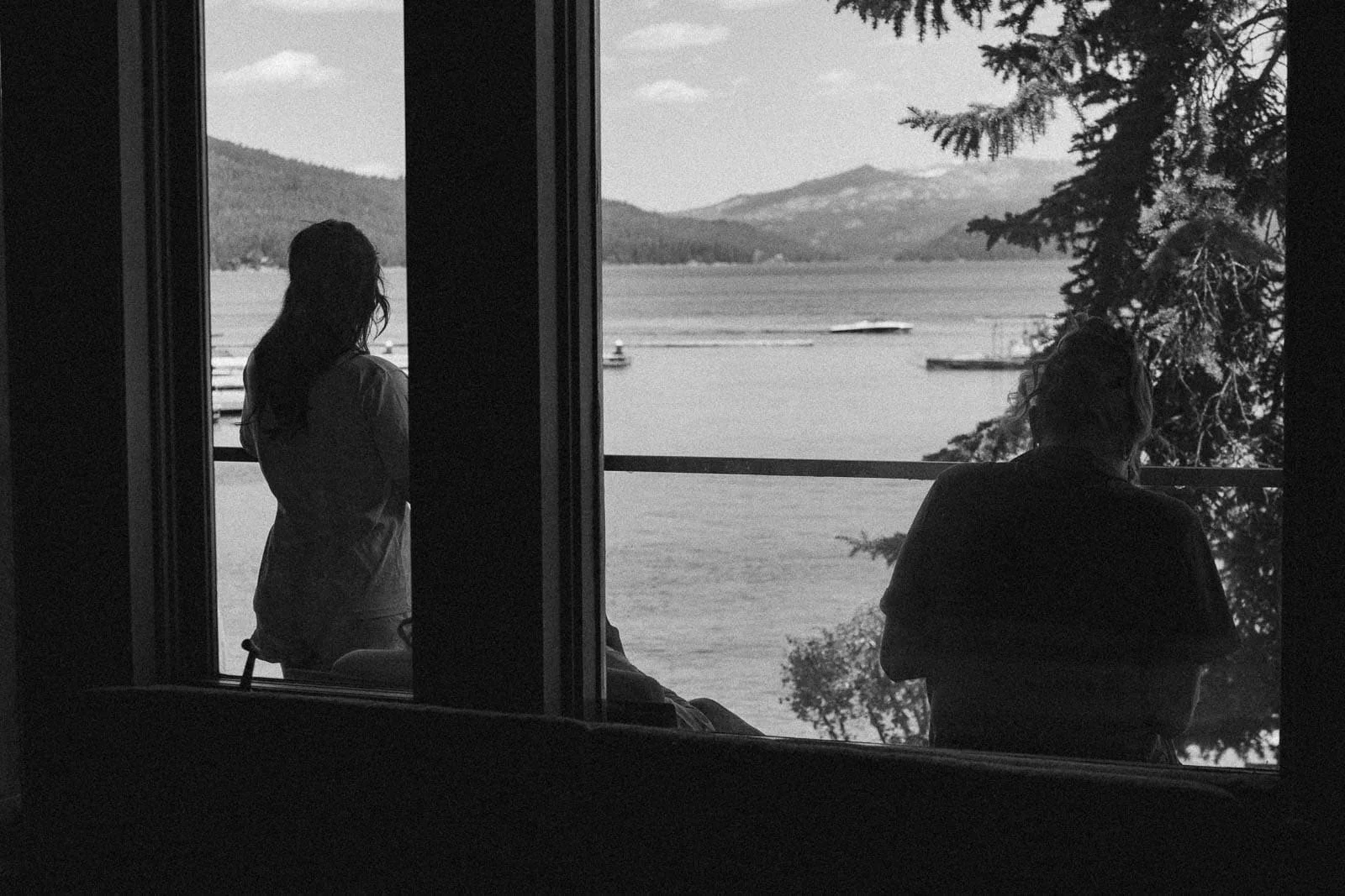  Two people are seen in silhouette inside a building, looking out a window at a lake with boats and mountains in the distance. Trees frame the scenic outdoor view under a bright sky. 