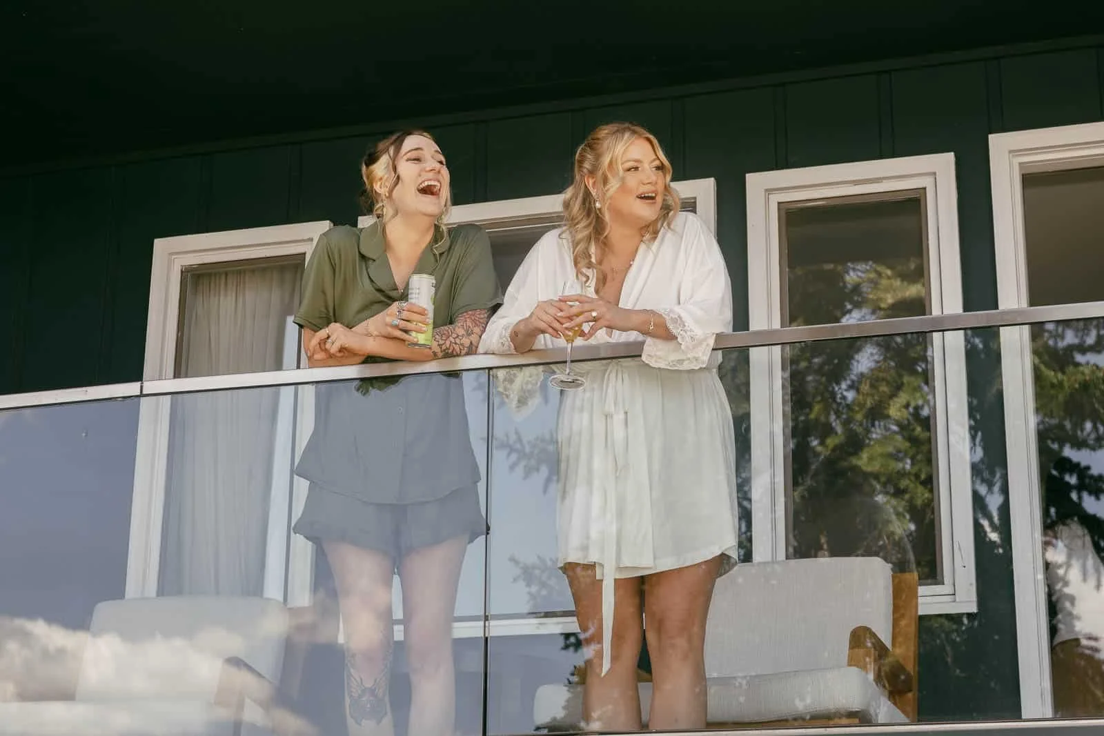  Two women stand on a balcony, smiling and laughing while holding drinks. They are dressed in light, casual outfits. The background shows windows and greenery reflected in the glass railing. 