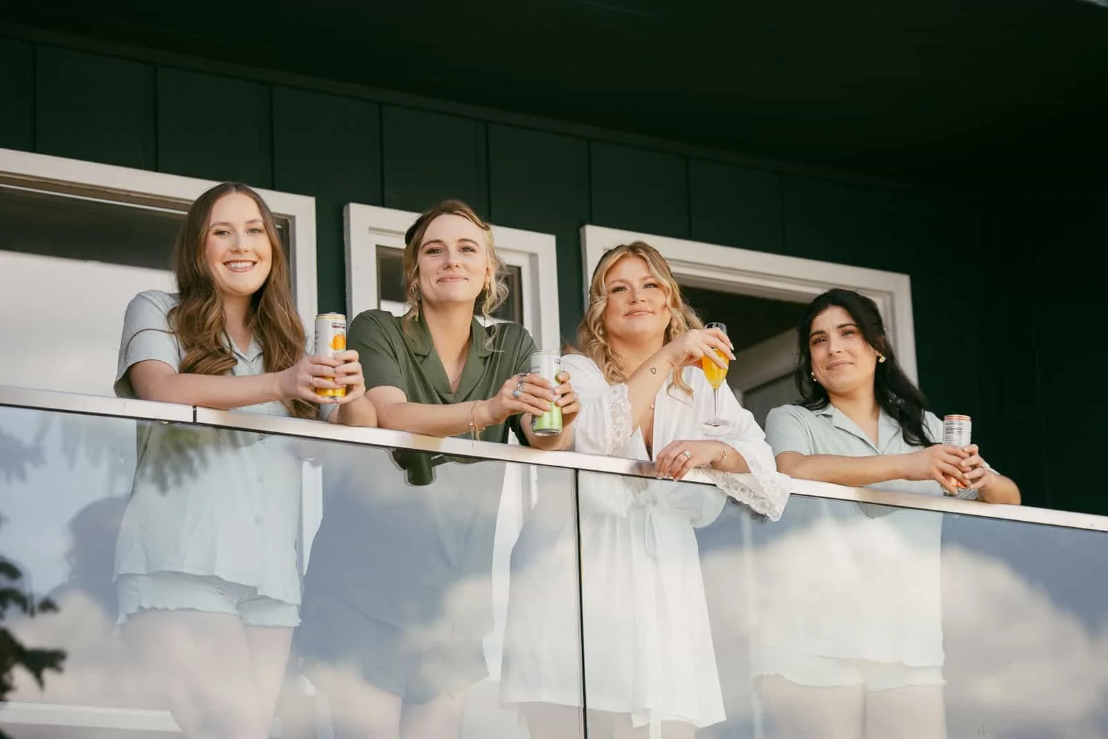  Four women in loungewear stand on a balcony, smiling and holding canned drinks, with windows and dark walls behind them. 