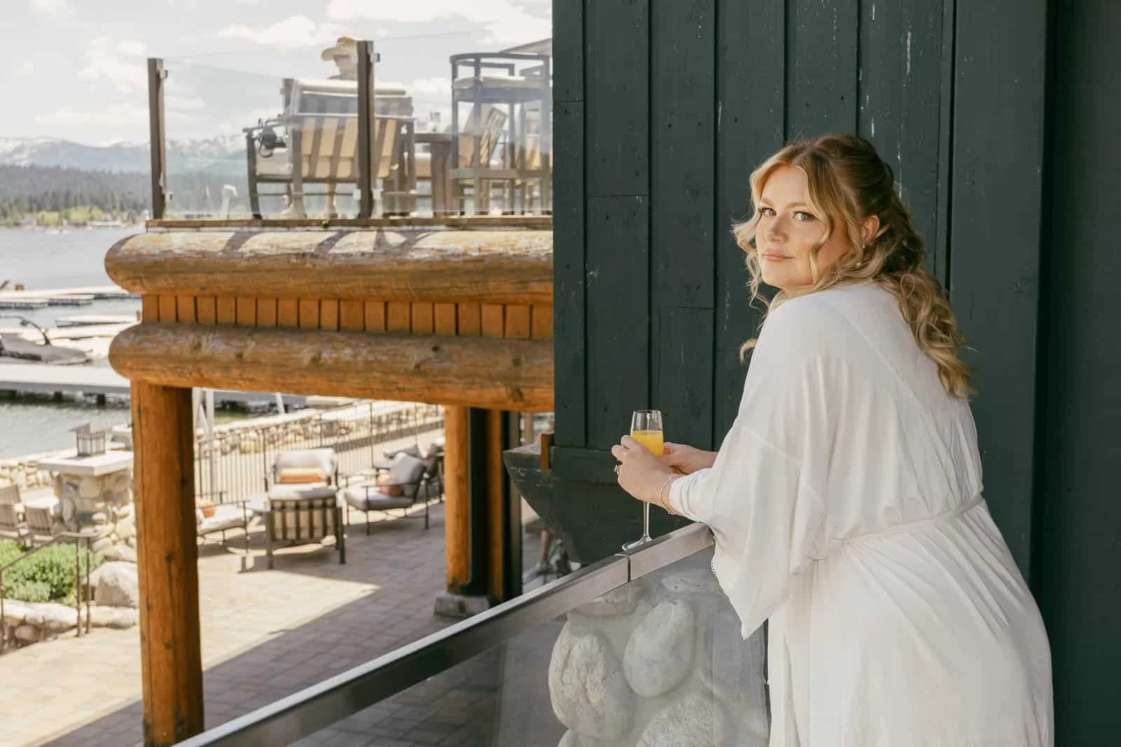  A bride in a white robe with wavy blonde hair holds a drink and leans on a balcony railing, gazing at the camera. Outdoor furniture and a lake are visible in the background. 