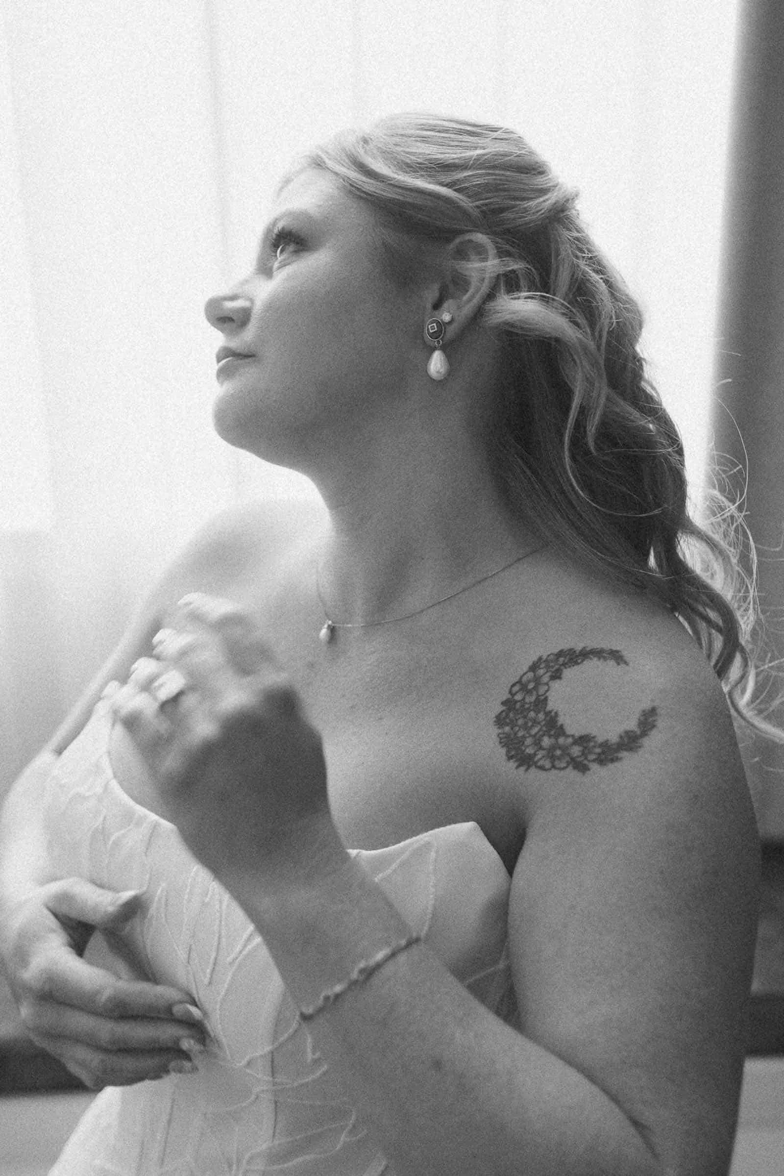  A bride in a strapless wedding dress looks up thoughtfully. She has wavy hair, wearing earrings, and a necklace. A crescent moon tattoo made of flowers is on her shoulder. The image is in black and white. 