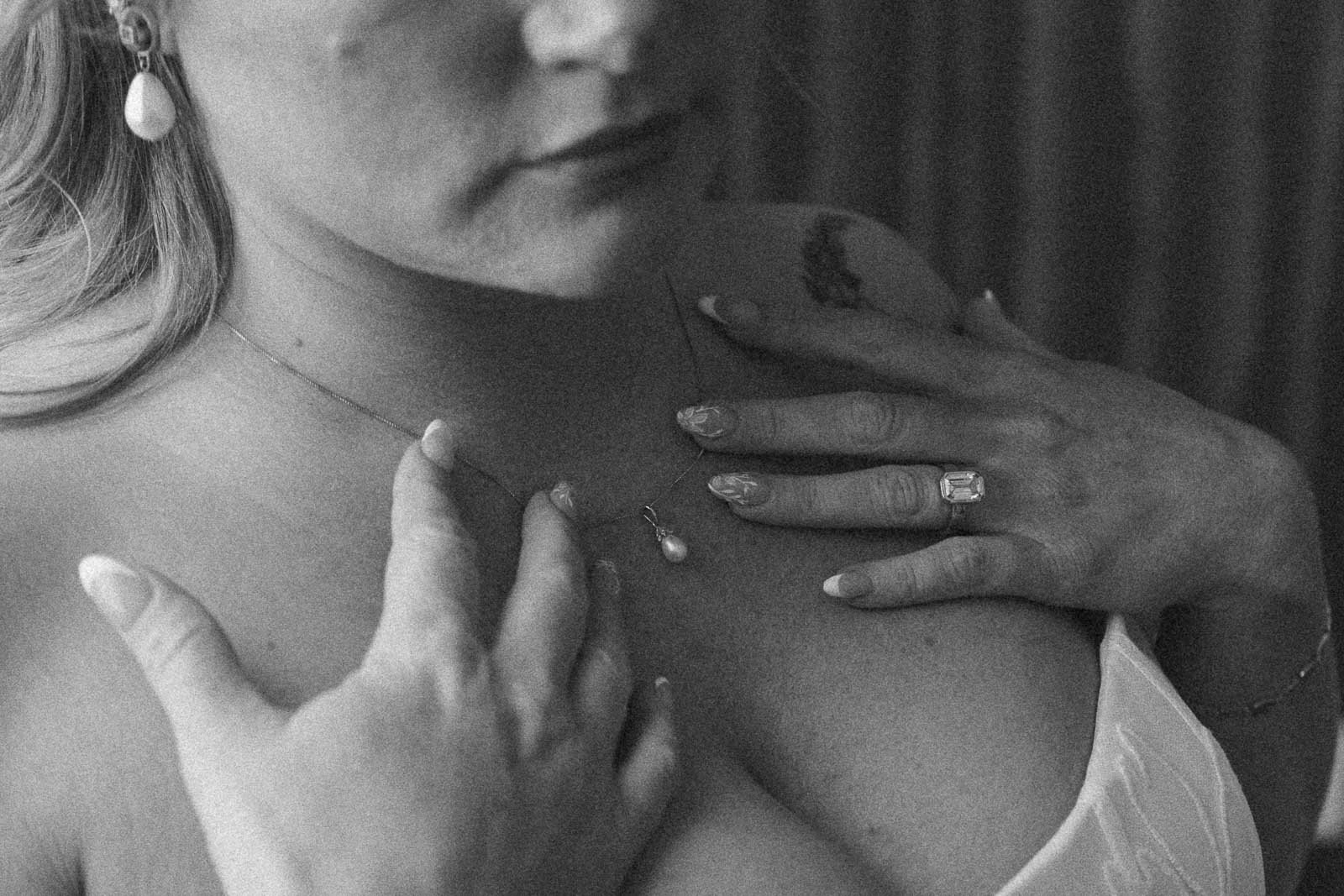  A bride in a strapless wedding dress touches her chest, showing a pearl necklace, pearl earring, and a ring on her finger. The image is in black and white and slightly out of focus. 