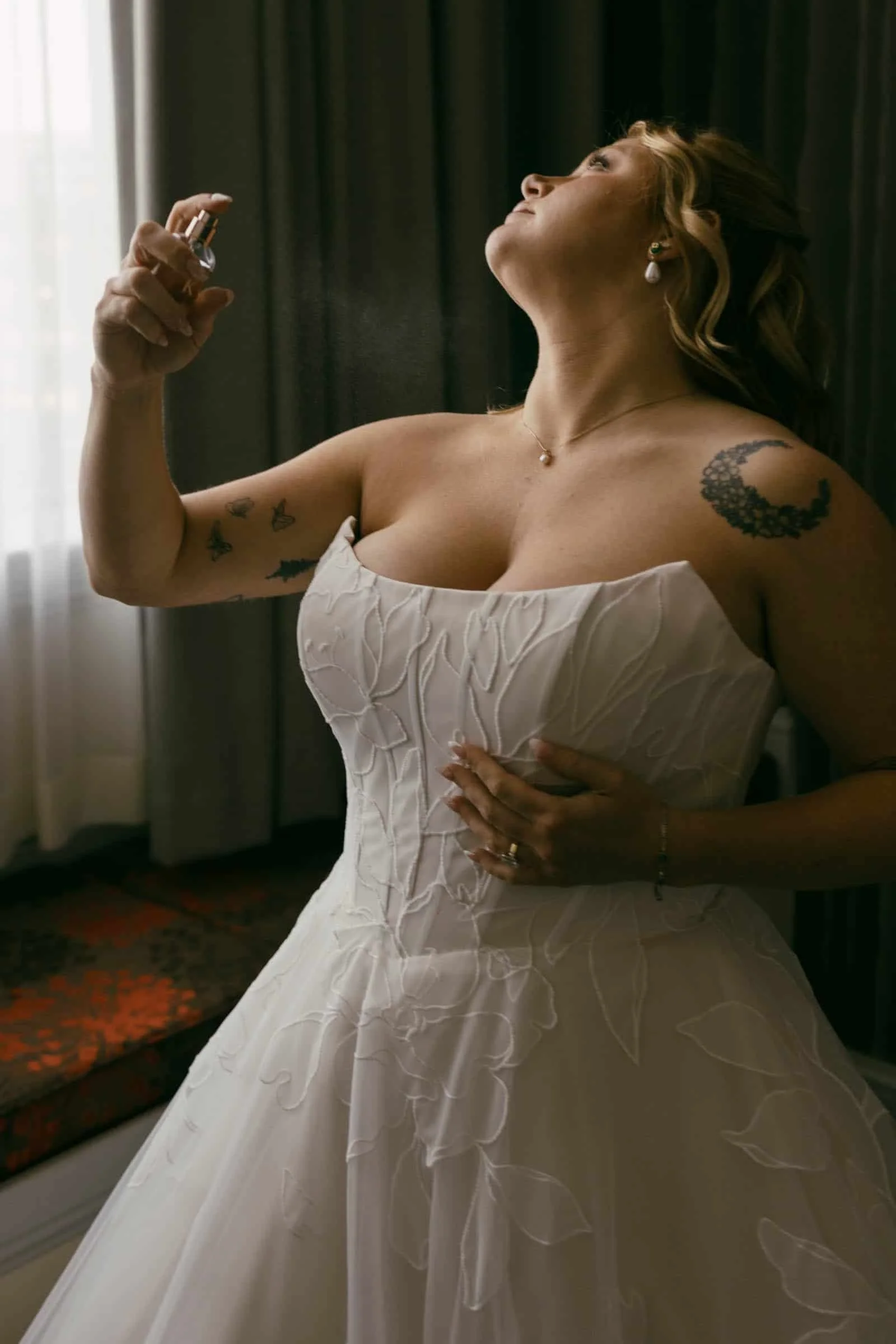  A bride in a white strapless gown sprays perfume on her neck while standing by a window with dark curtains. 