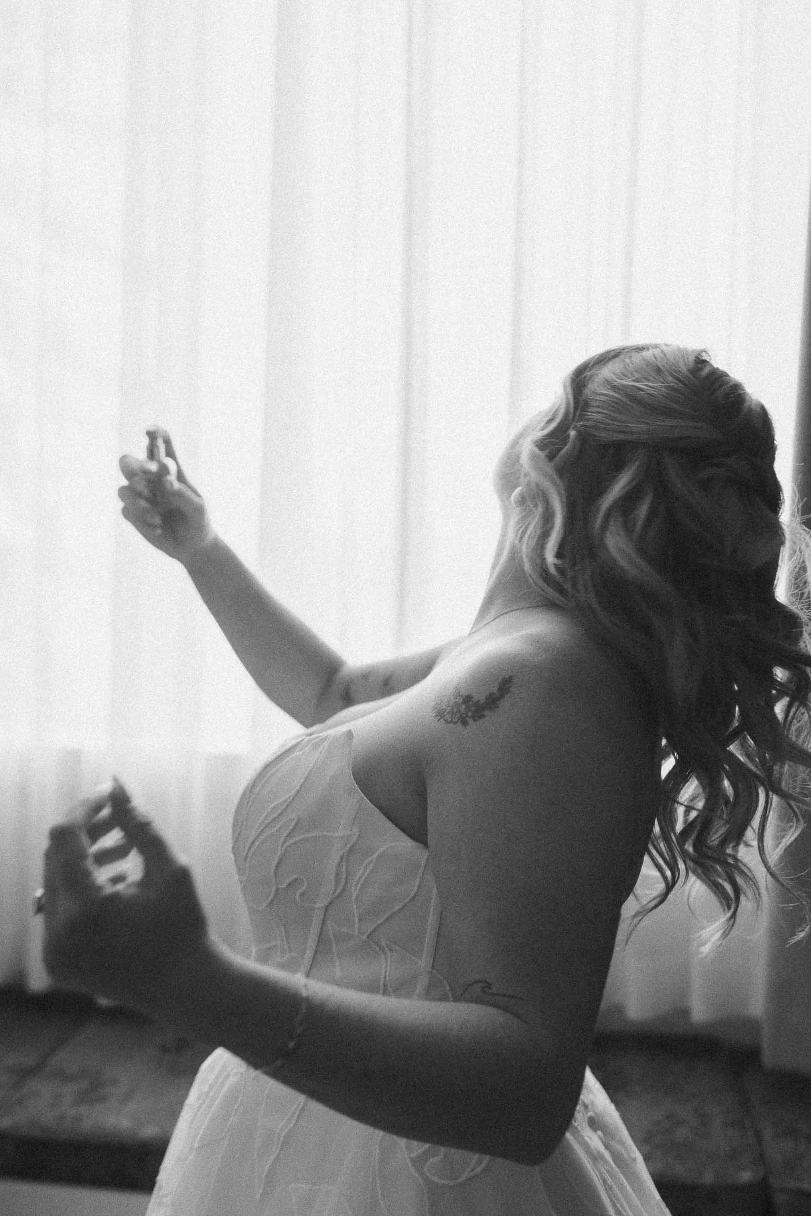  A gride in a white dress with wavy hair and tattoos sprays perfume near a window with sheer curtains, backlit by natural light. 