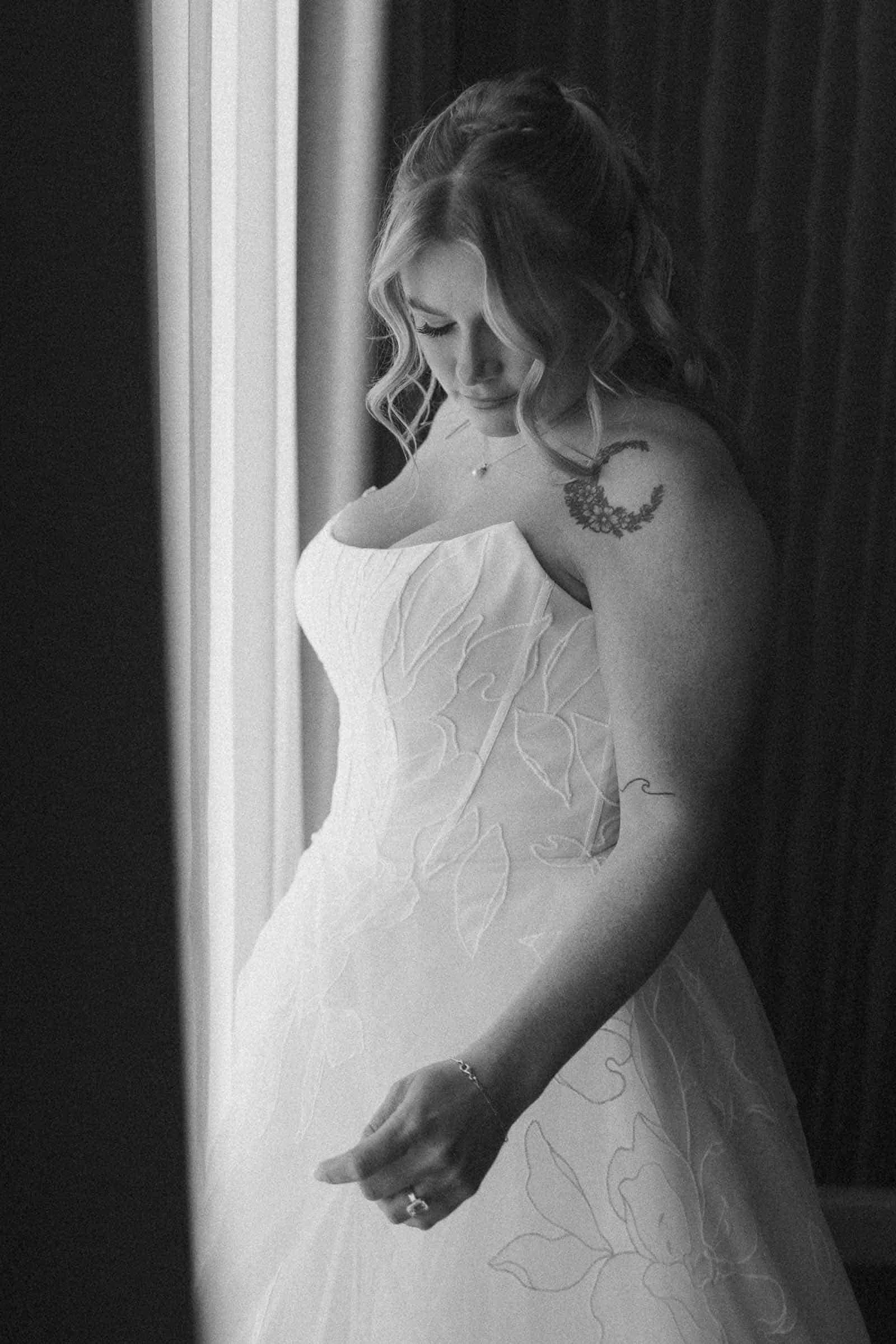  A woman in a strapless wedding dress stands by a window, looking down. Her wavy hair is partially pulled back, and she has a floral tattoo on her shoulder. The image is in black and white. 
