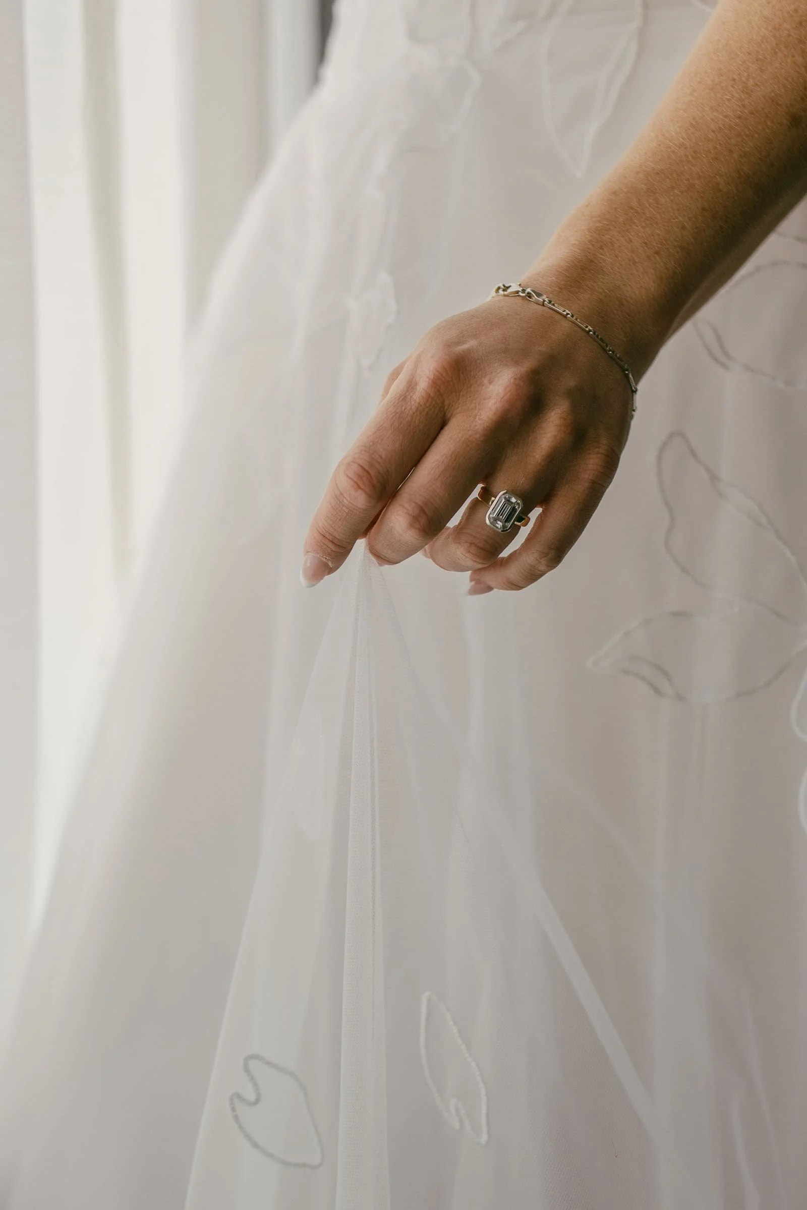  A close-up of a hand holding a white, sheer wedding dress with embroidered leaf patterns. The hand wears a silver ring with a large rectangular gemstone and a delicate bracelet. 