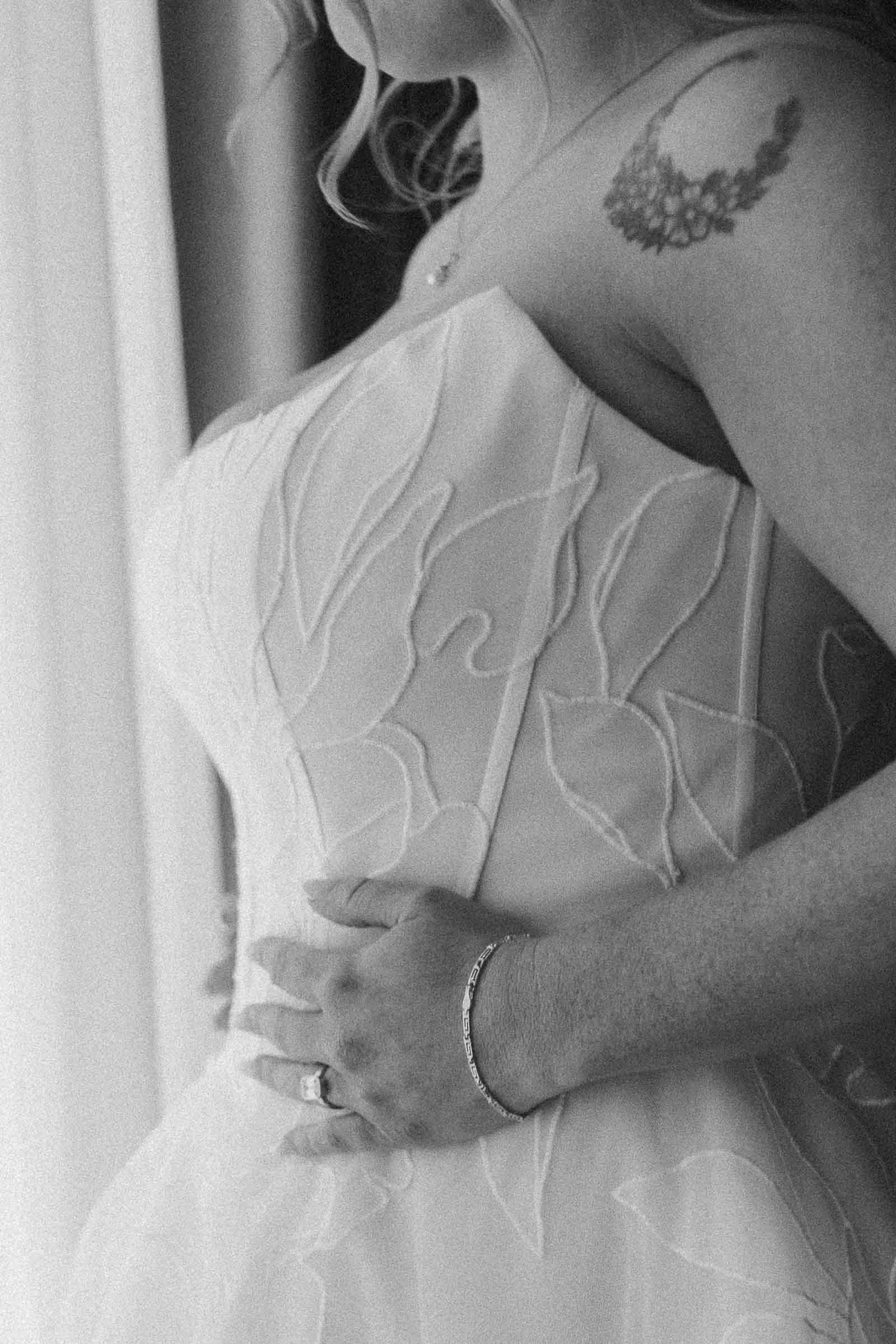  A woman in a strapless wedding dress with leaf patterns stands by a window. She wears a bracelet and ring, and has a floral tattoo on her shoulder. 