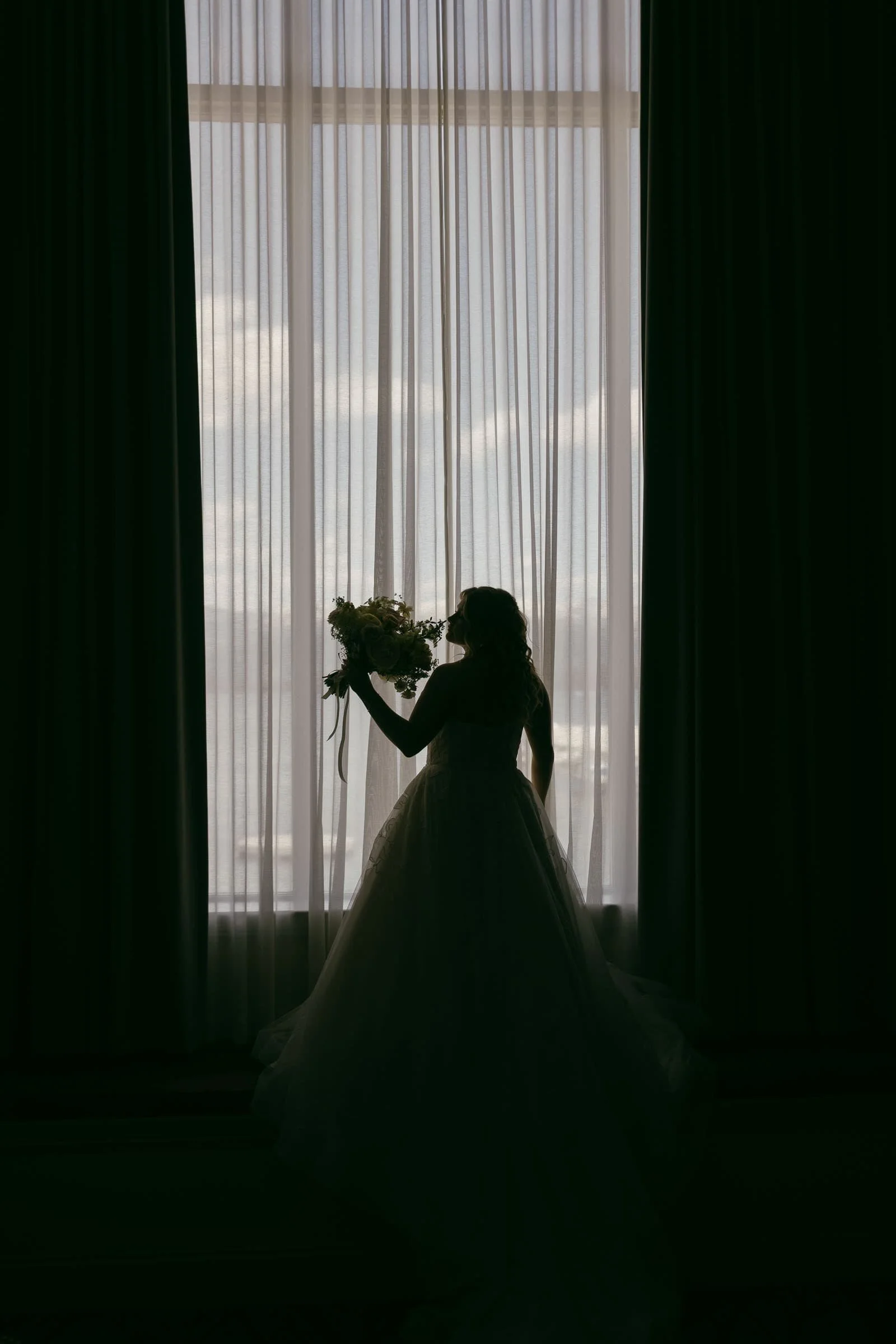  A bride in a gown holds a bouquet and stands silhouetted against a large window with sheer curtains, softly lit by daylight outside. 