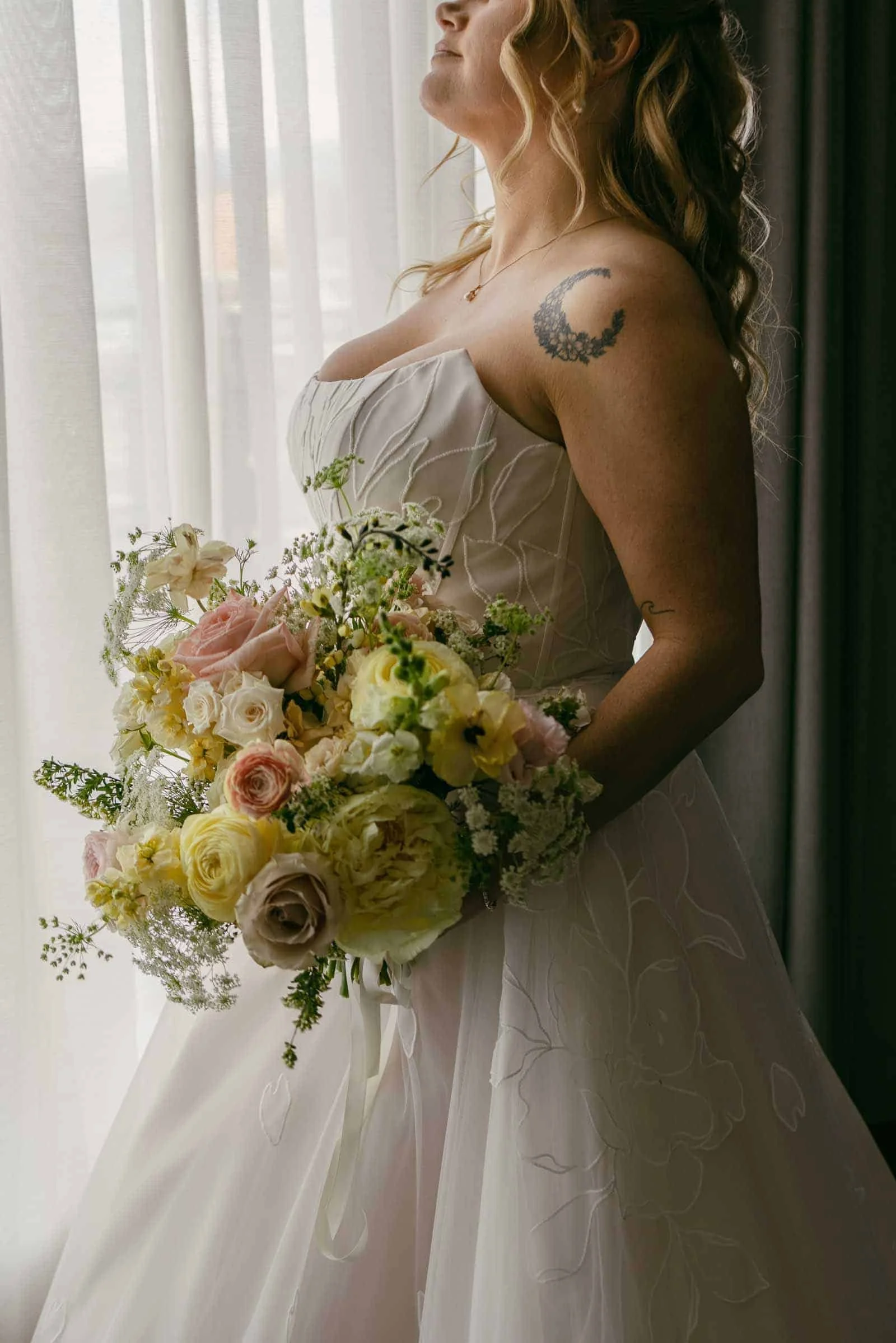  A bride in a white dress holds a bouquet of pastel flowers by a window. She has a crescent moon tattoo on her shoulder and soft curls in her hair. Light streams through sheer curtains behind her. 