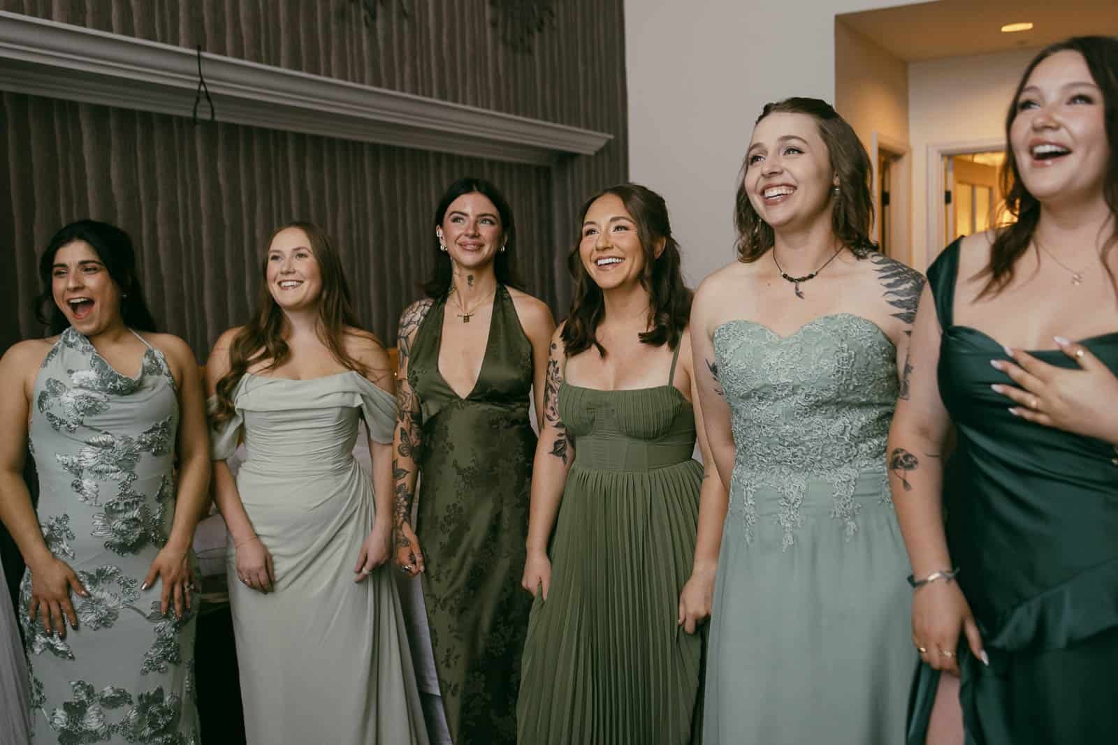  Six women in coordinating green dresses stand indoors, smiling and looking excited, possibly reacting to something off-camera at a formal event. 