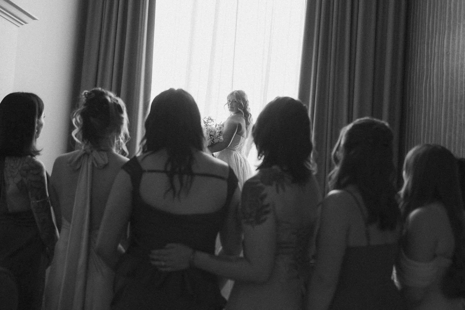  A bride stands in front of a bright window, holding a bouquet, while six bridesmaids in dresses stand together in the foreground, facing her. The photo is in black and white. 