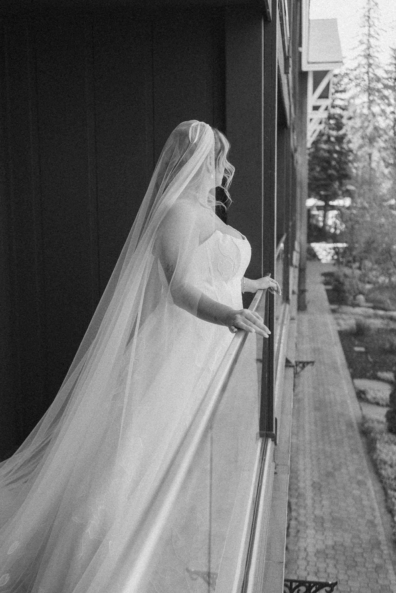  A bride in a wedding dress and veil stands on a balcony, gazing out over a garden path. The photo is black and white and captures a serene, candid moment from the side. 