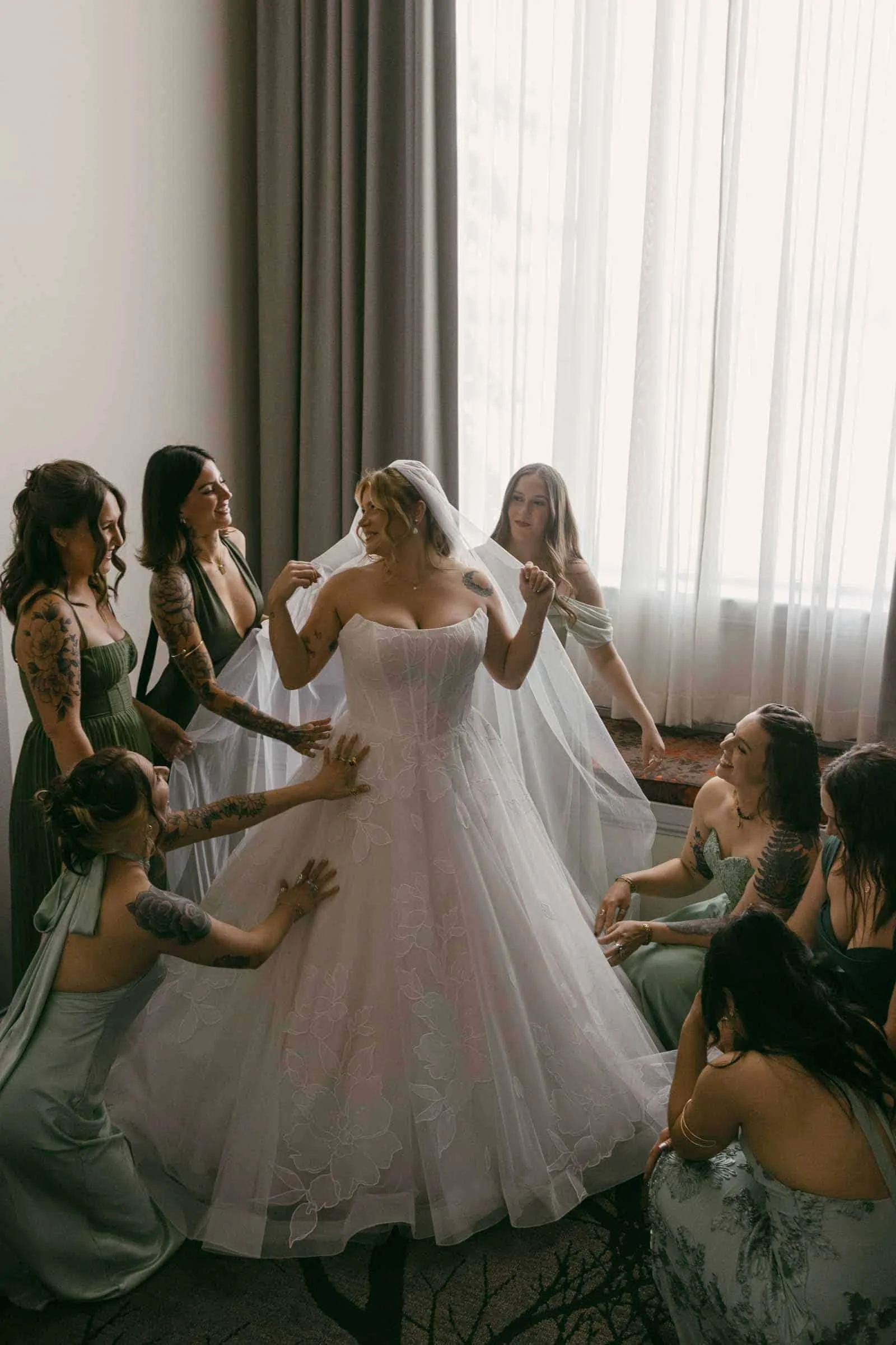  A bride in a strapless gown stands surrounded by bridesmaids in pale green dresses, all smiling and helping her adjust her dress near a window with sheer curtains. 