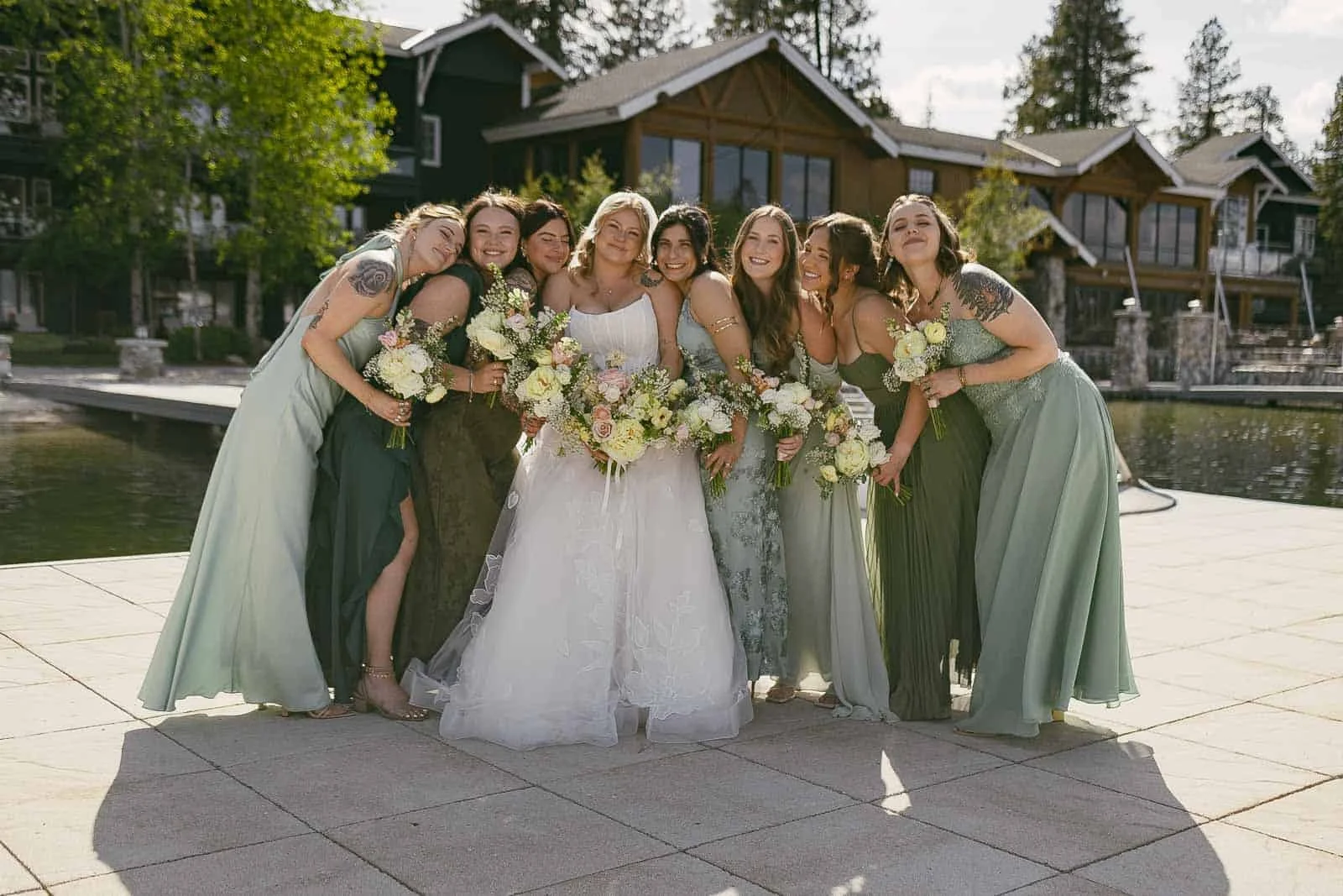 A bride in a white dress stands outdoors, surrounded by seven bridesmaids in green dresses, all smiling and holding bouquets, with rustic buildings and water in the background. 