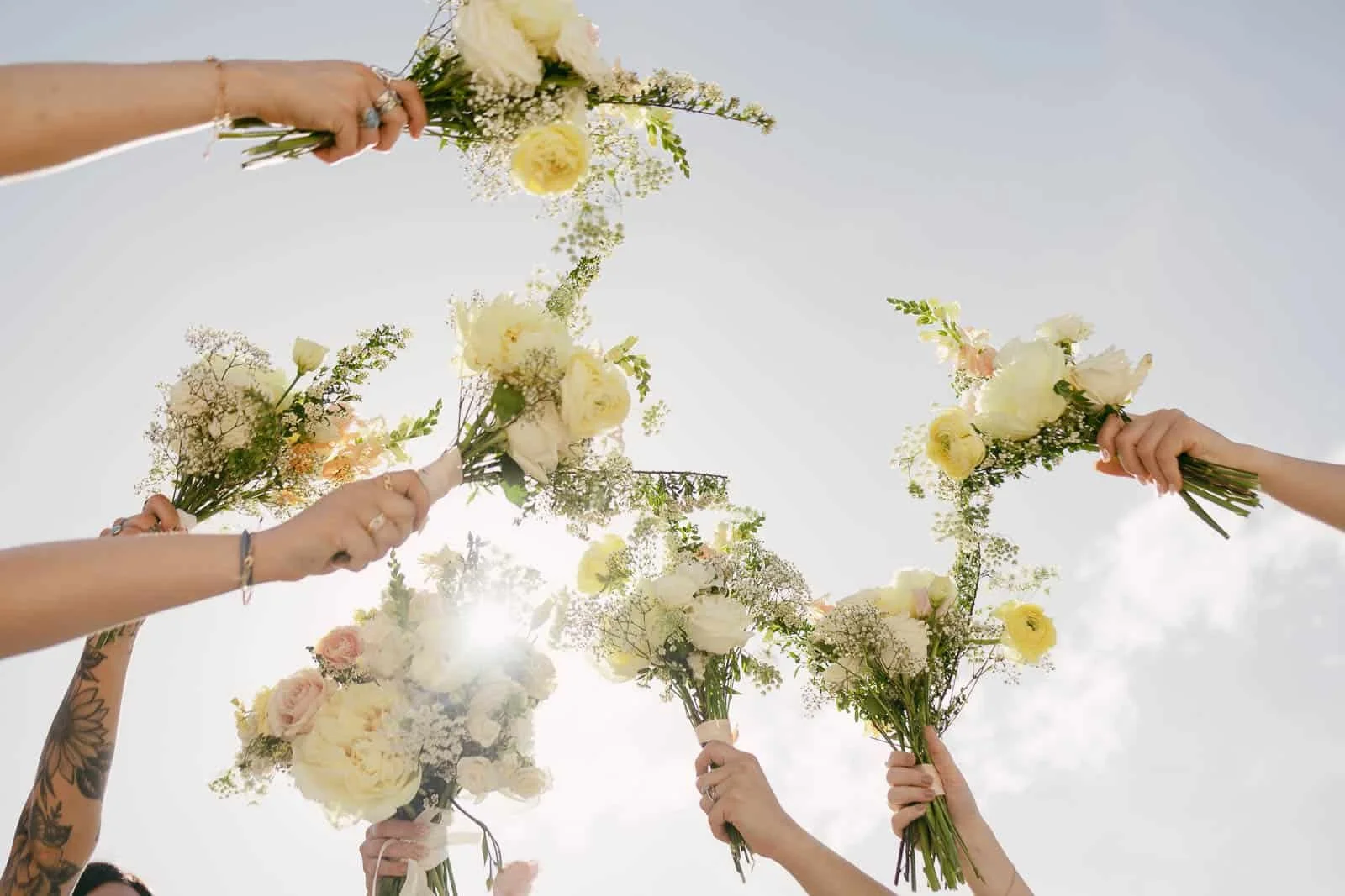  Several hands hold bouquets of white and yellow flowers up into a bright, sunny sky, with sunlight shining through and a few clouds visible in the background. 