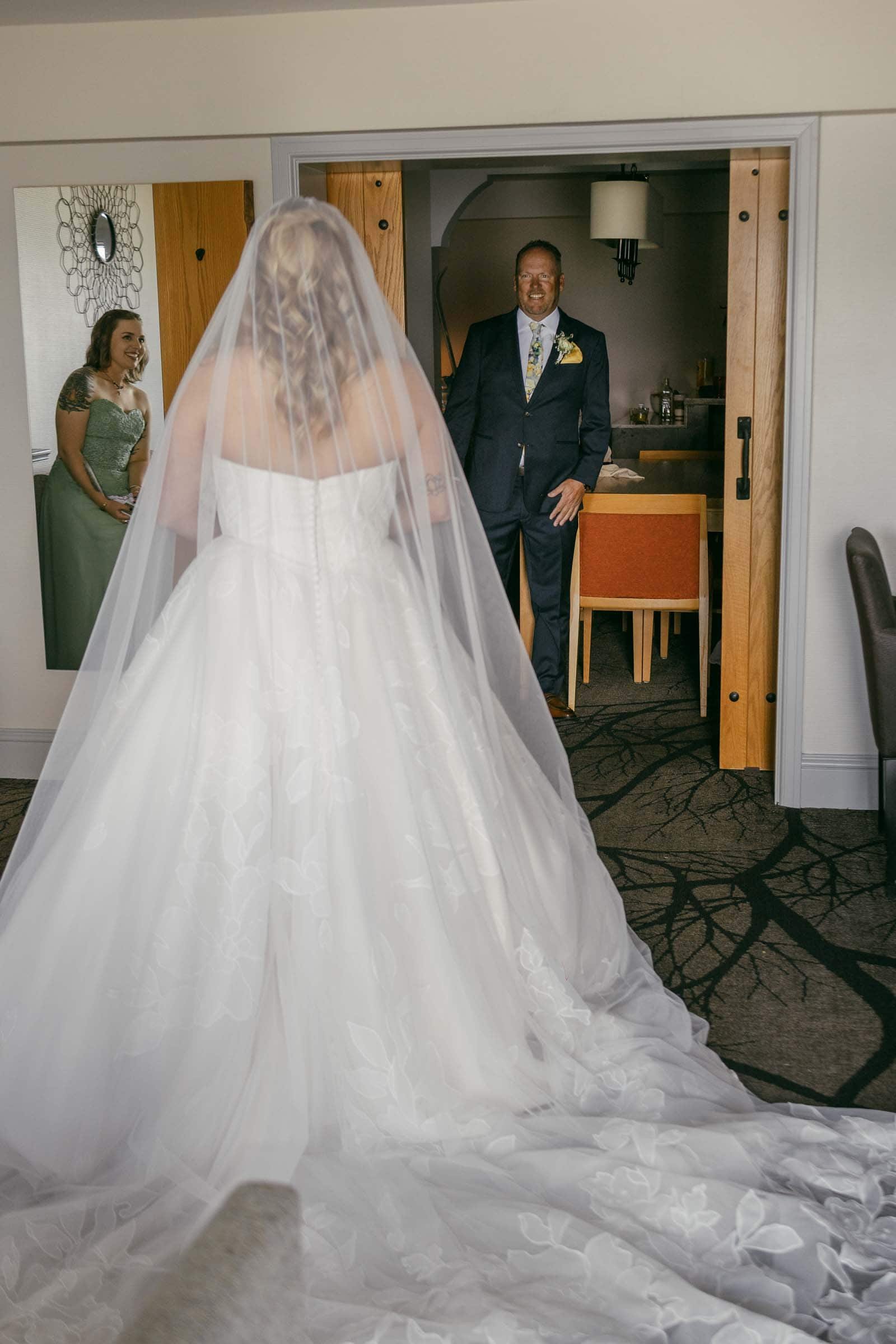  A bride in a white gown faces her dad in a suit, who smiles warmly. A woman in a green dress watches from the left, also smiling. They are indoors, near a table and chairs, with wooden doors in the background. 