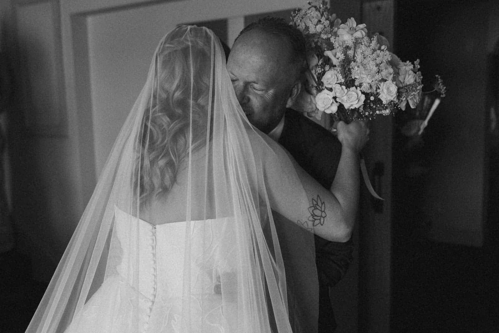  A bride in a veil hugs her dad, who smiles with his eyes closed, holding a bouquet of flowers. The moment appears emotional and joyful, captured indoors in black and white. They have just done a first look 