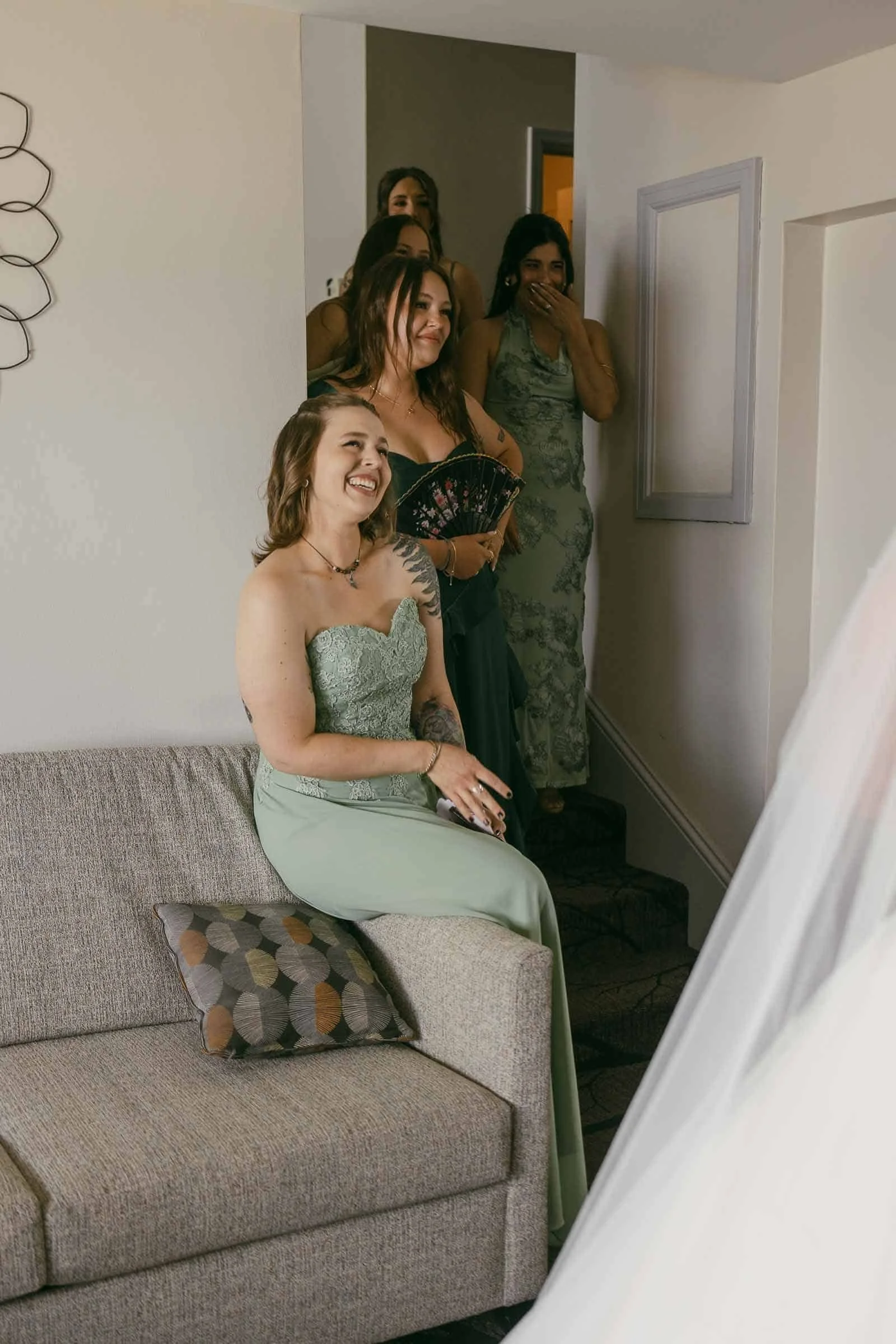  A group of bridesmaids in green dresses smile and look emotional as they see the bride in her gown, who is just out of frame. One bridesmaid sits on a couch, while others stand on a staircase behind her. 