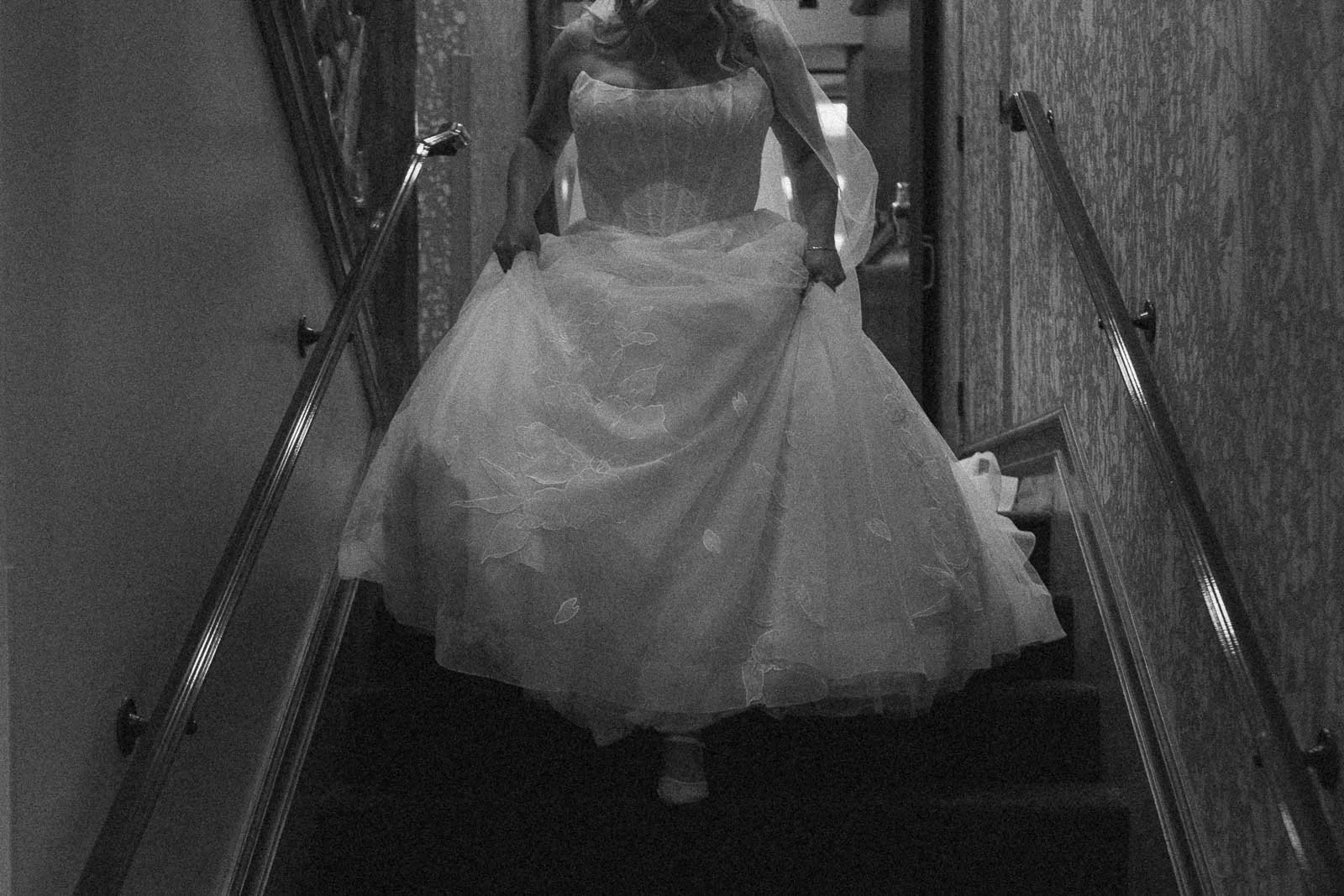  A person in a wedding dress descends a carpeted staircase, holding up the skirt of the dress. The image is in black and white and appears to be candid. 