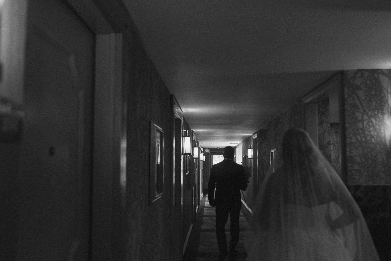  A bride in a wedding dress and veil walks down a dimly lit hallway behind a man in a suit. The scene is in black and white, with light coming from wall lamps. 