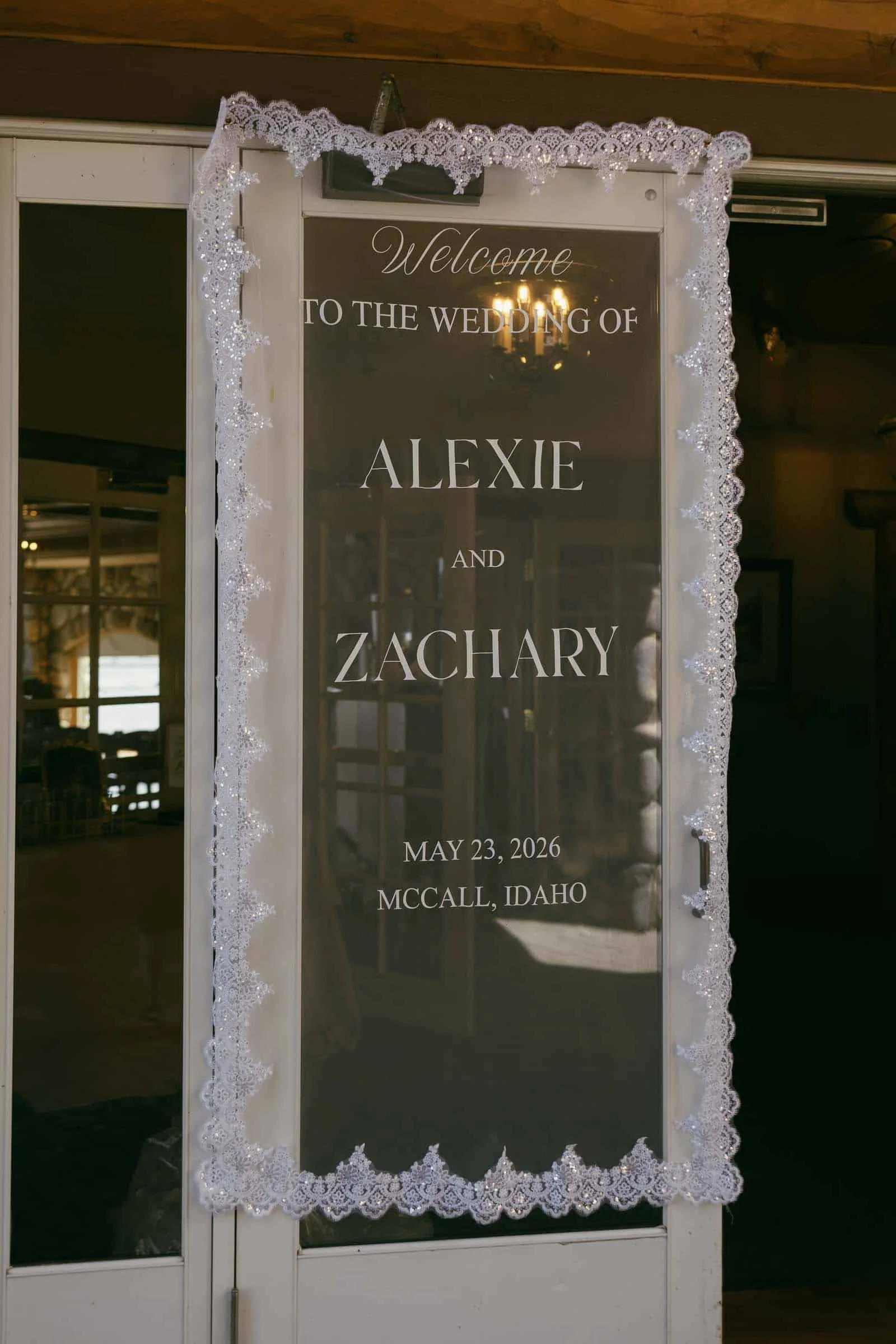  A translucent wedding sign with lace trim reads, "Welcome to the wedding of Alexie and Zachary, May 23, 2026, McCall, Idaho," and is displayed on glass doors. 