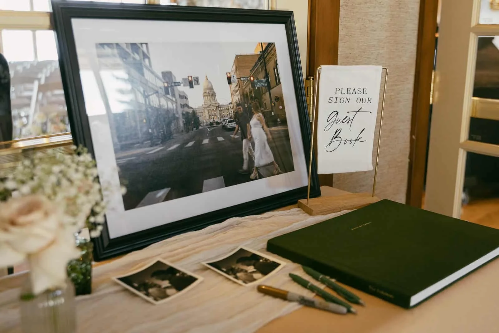  Wedding guest book table with framed photo of a couple crossing a street, a sign reading “Please sign our Guest Book,” flowers, two pens, and black-and-white photos. 