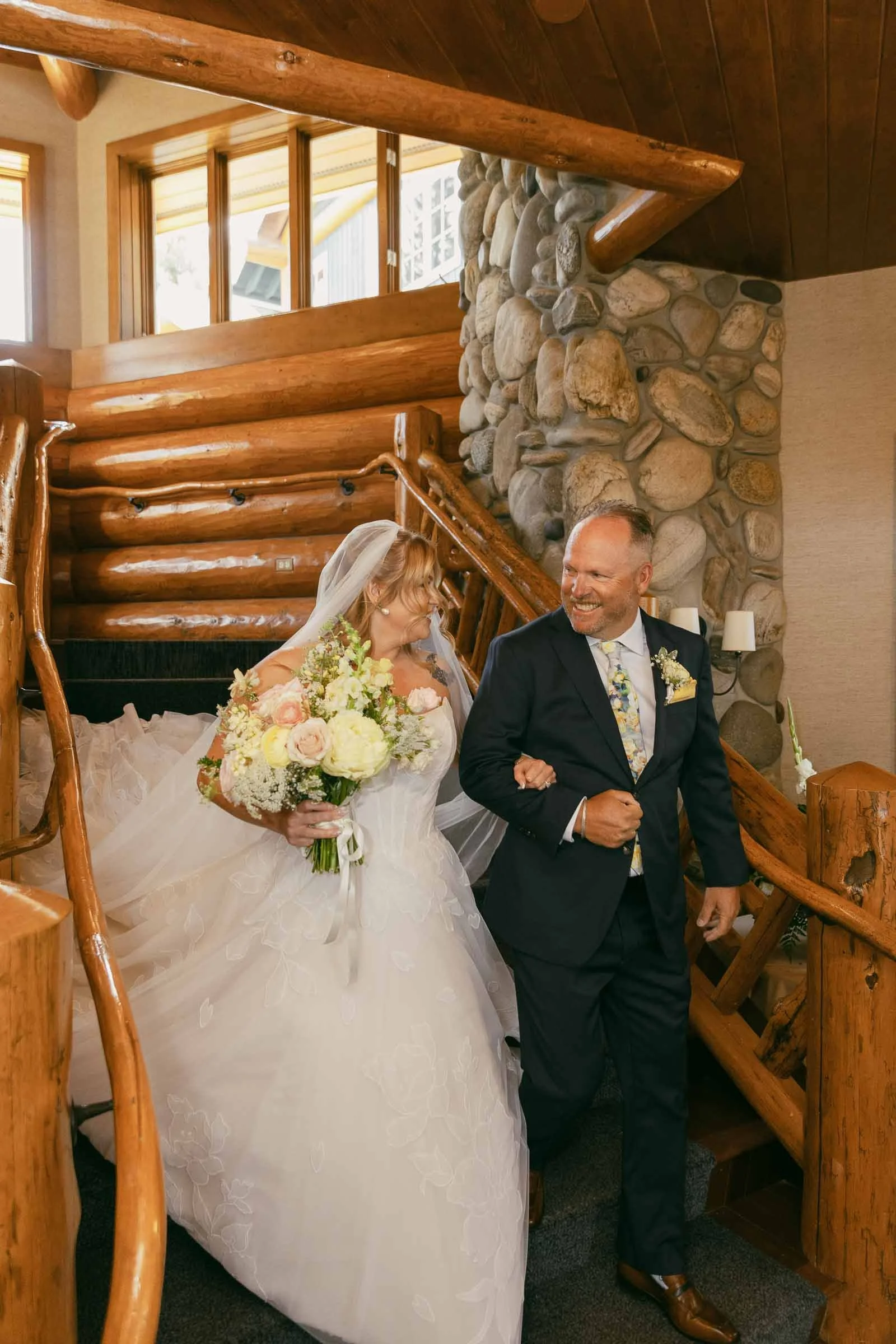  A bride in a white gown holding a bouquet walks down wooden stairs with a smiling man in a suit at her side inside a rustic, log cabin-style venue. 