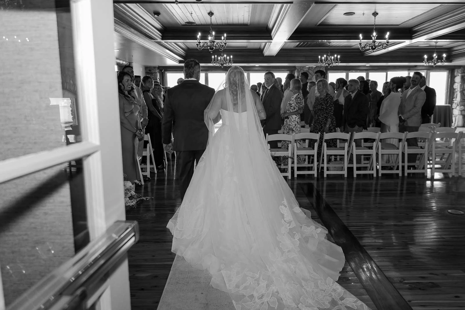  A bride in a flowing gown walks down the aisle with a man, likely her father, in a room filled with standing guests. The scene is captured in black and white. 