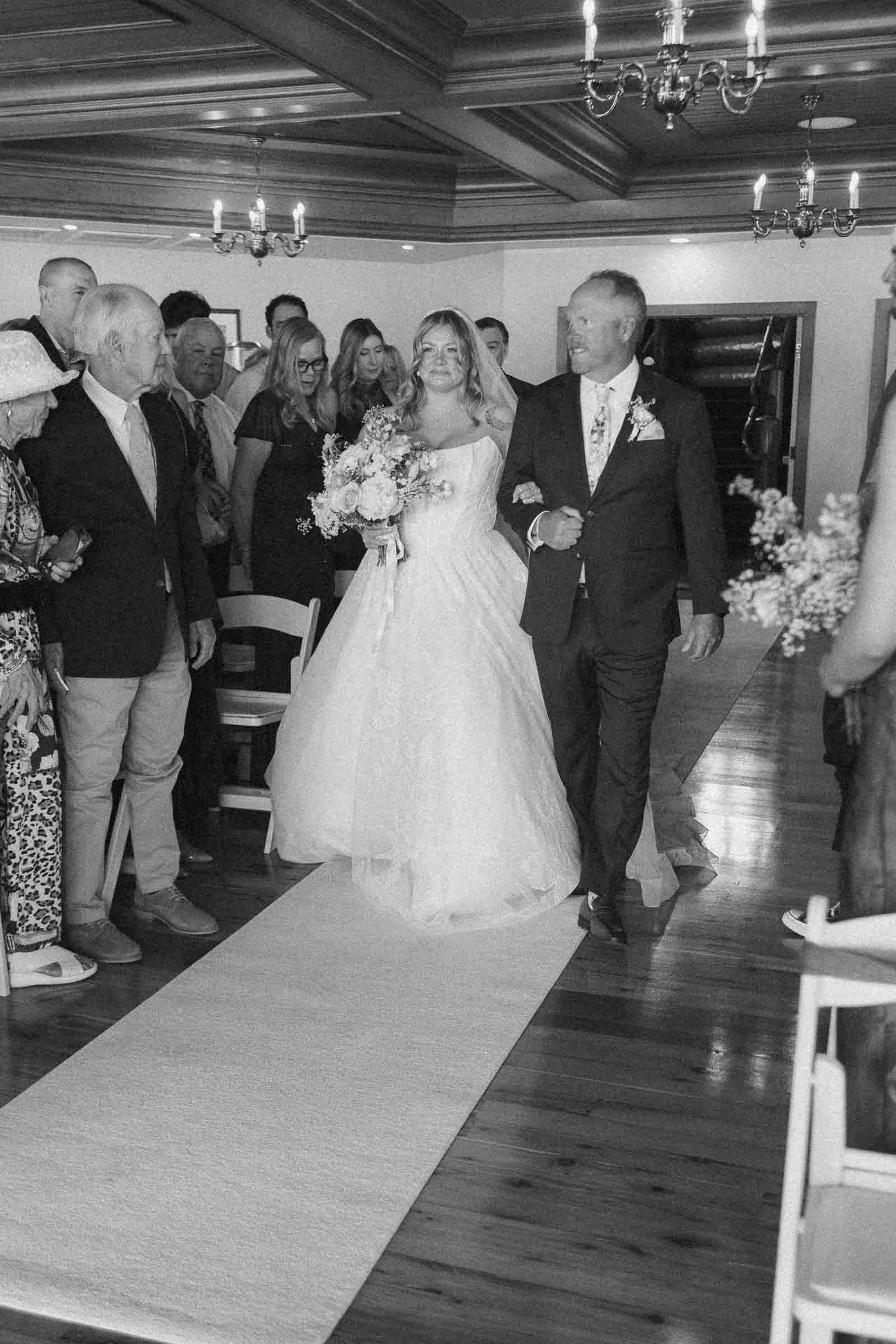  A bride in a wedding dress walks down the aisle on a white runner, accompanied by an older man in a suit, while guests watch and smile in a warmly lit indoor venue. 