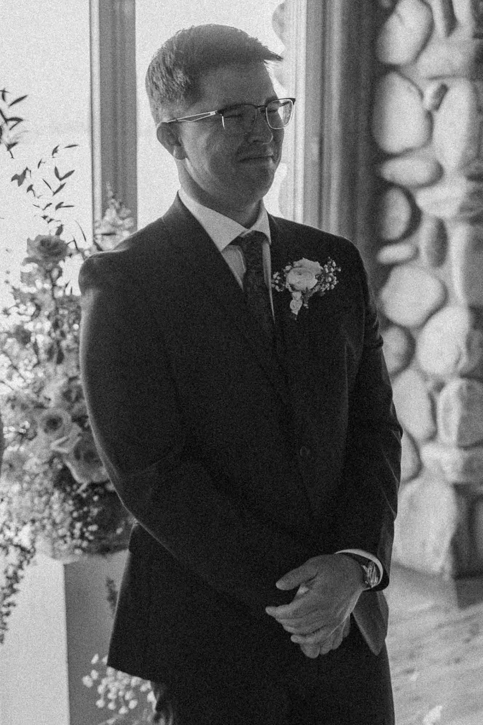  A man in a suit with a boutonniere stands indoors, smiling softly. The background features a stone wall and floral arrangements. The image is in black and white. 