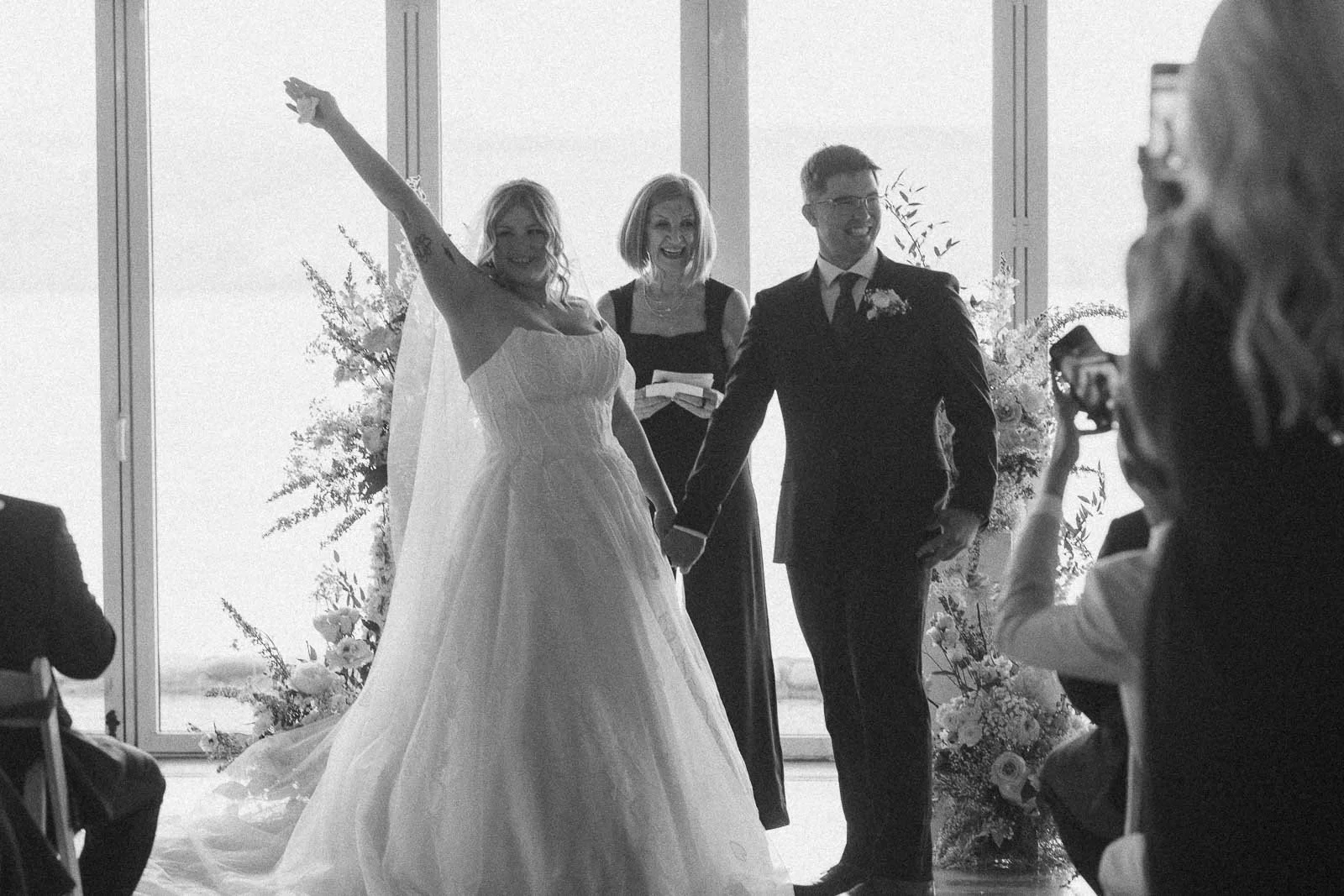  A bride and groom stand holding hands at their wedding ceremony, smiling with a woman officiant. The bride raises her arm in celebration. Guests are seated, and one person takes a photo. 