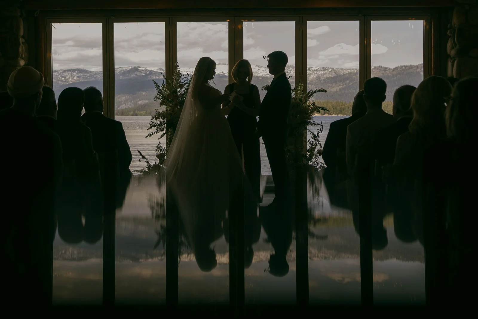  A bride and groom stand with an officiant by large windows overlooking a lake and mountains, silhouetted against natural light, with guests seated in shadow. 
